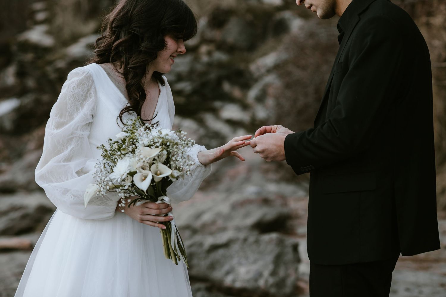 A bride in a long-sleeve white dress holds a white floral bouquet while exchanging rings during an outdoor ceremony.