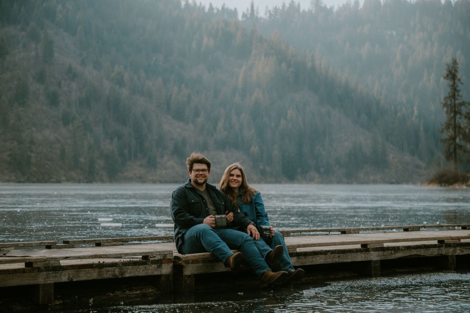 Couple sitting together on a wooden dock by a misty lake surrounded by evergreen forest.