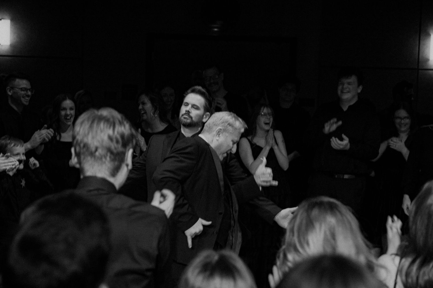 Two men embrace on a dimly lit stage surrounded by applauding guests at a formal evening event.