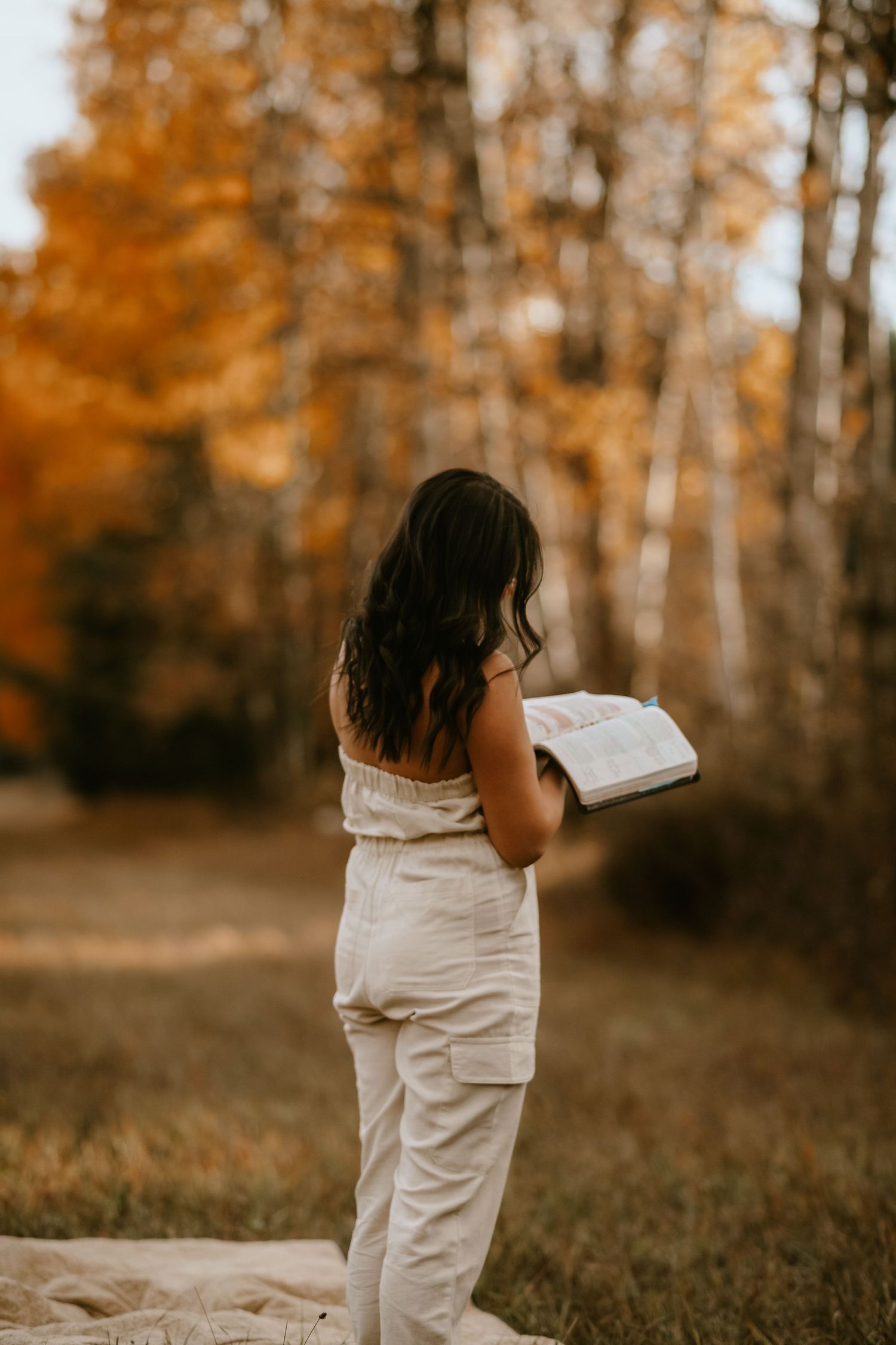 A person in white clothing reads a book outdoors during autumn with golden trees in the background.