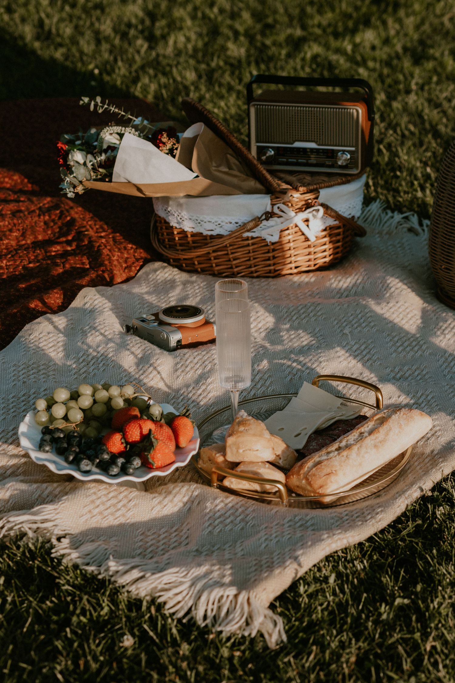 Vintage picnic setup on blanket with wicker basket, radio, fresh fruit, and sandwiches on sunny grass.