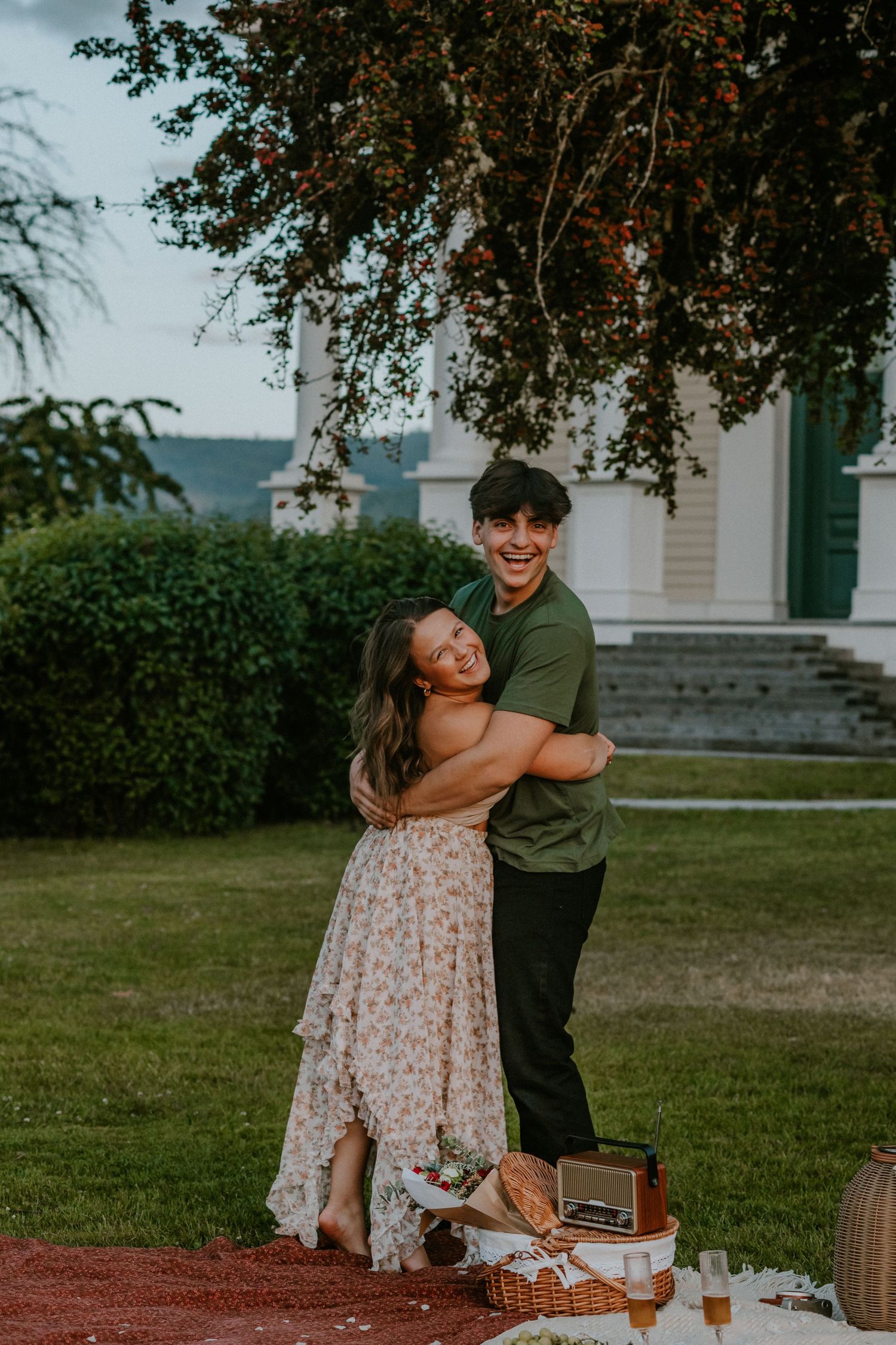 A couple embraces outdoors on a summer evening, standing near a picnic setup with blankets and decor under tree branches.