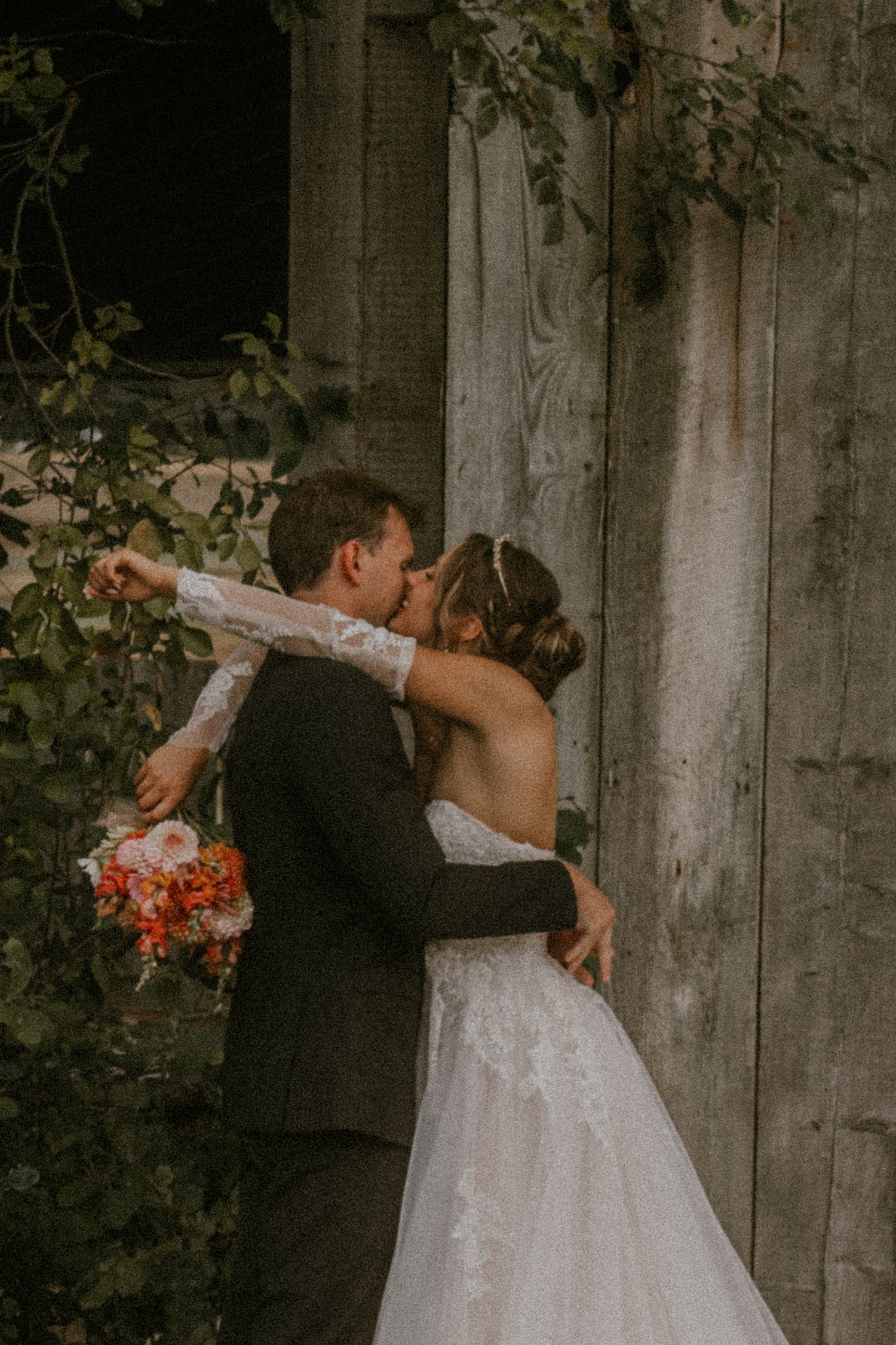 Bride and groom share a romantic kiss near a rustic barn door surrounded by lush greenery, bride holding a colorful bouquet.