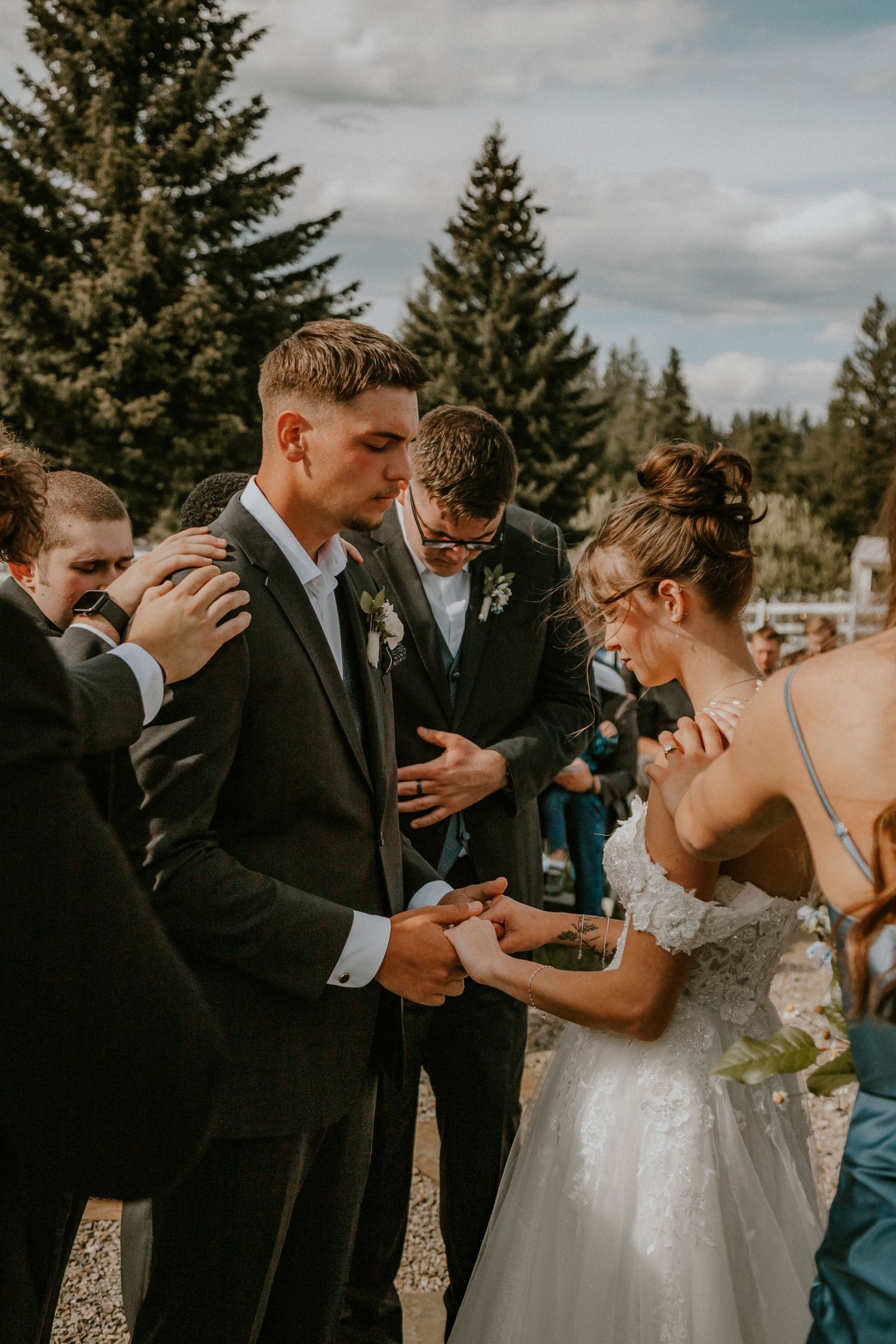 Emotional wedding party members embrace and show support for couple during outdoor ceremony with evergreen trees.
