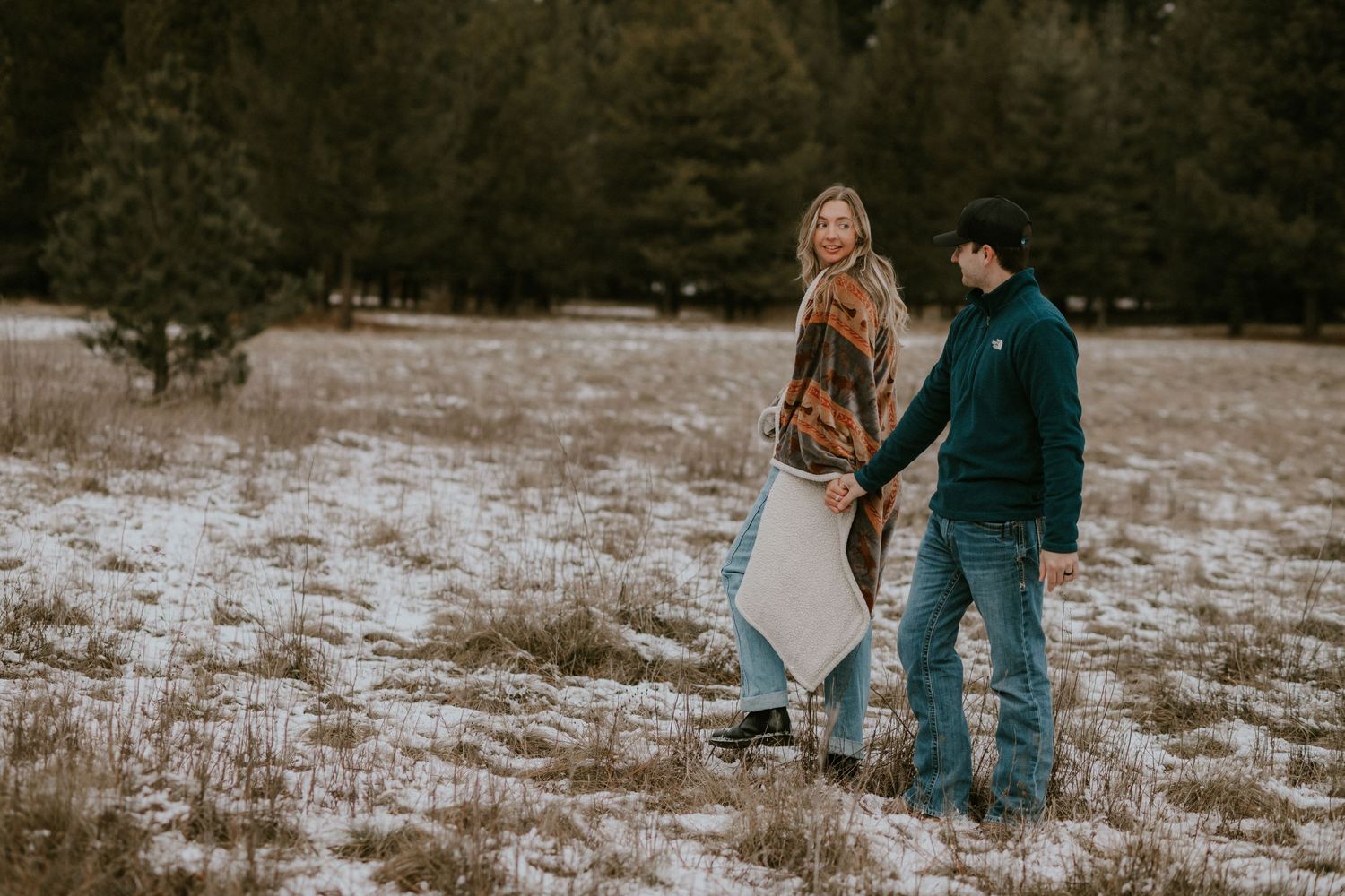 A couple walks hand in hand through a snowy winter field during a romantic outdoor engagement photo session.