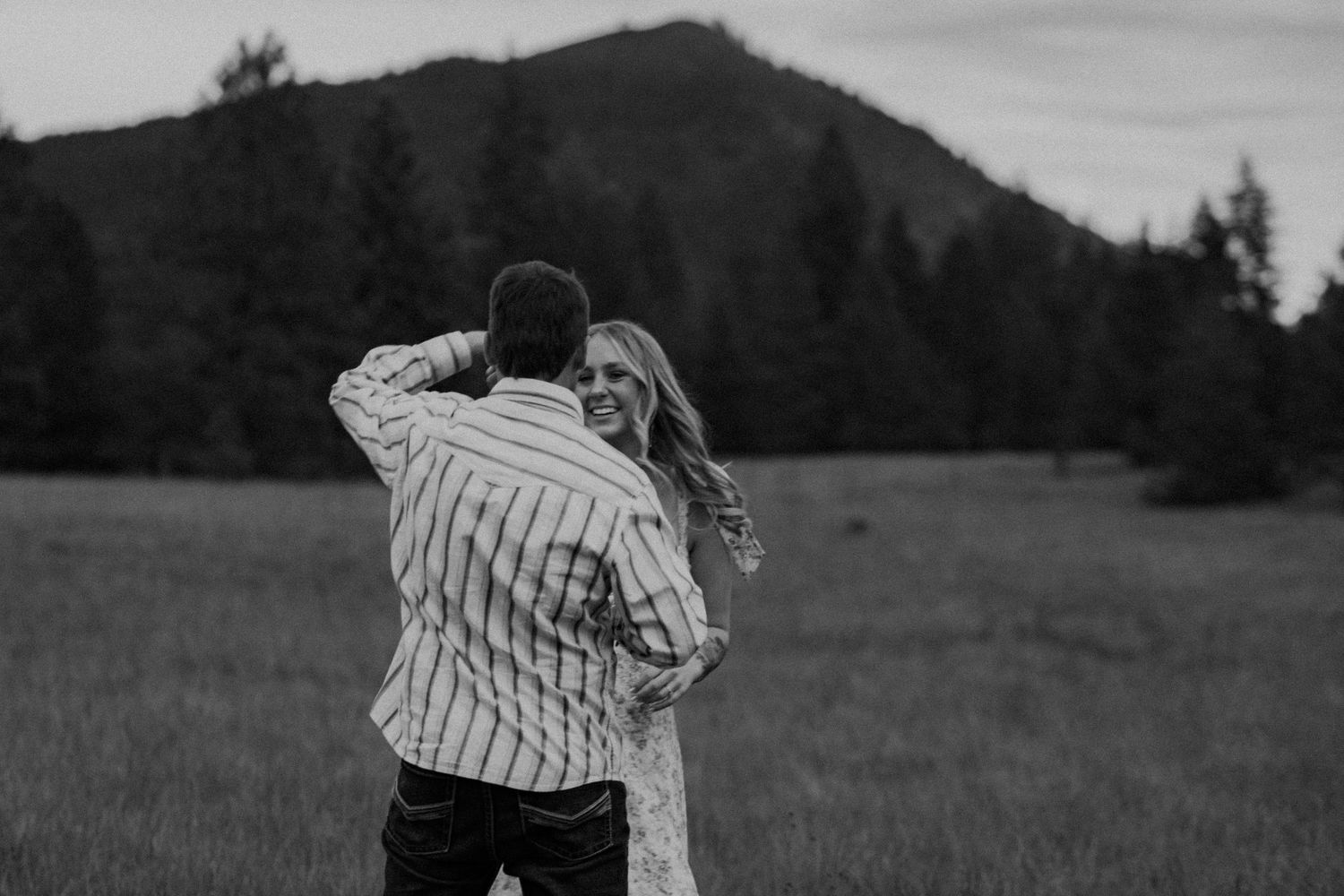 Couple embraces in a field at dusk with mountain silhouette and trees in black and white photography.