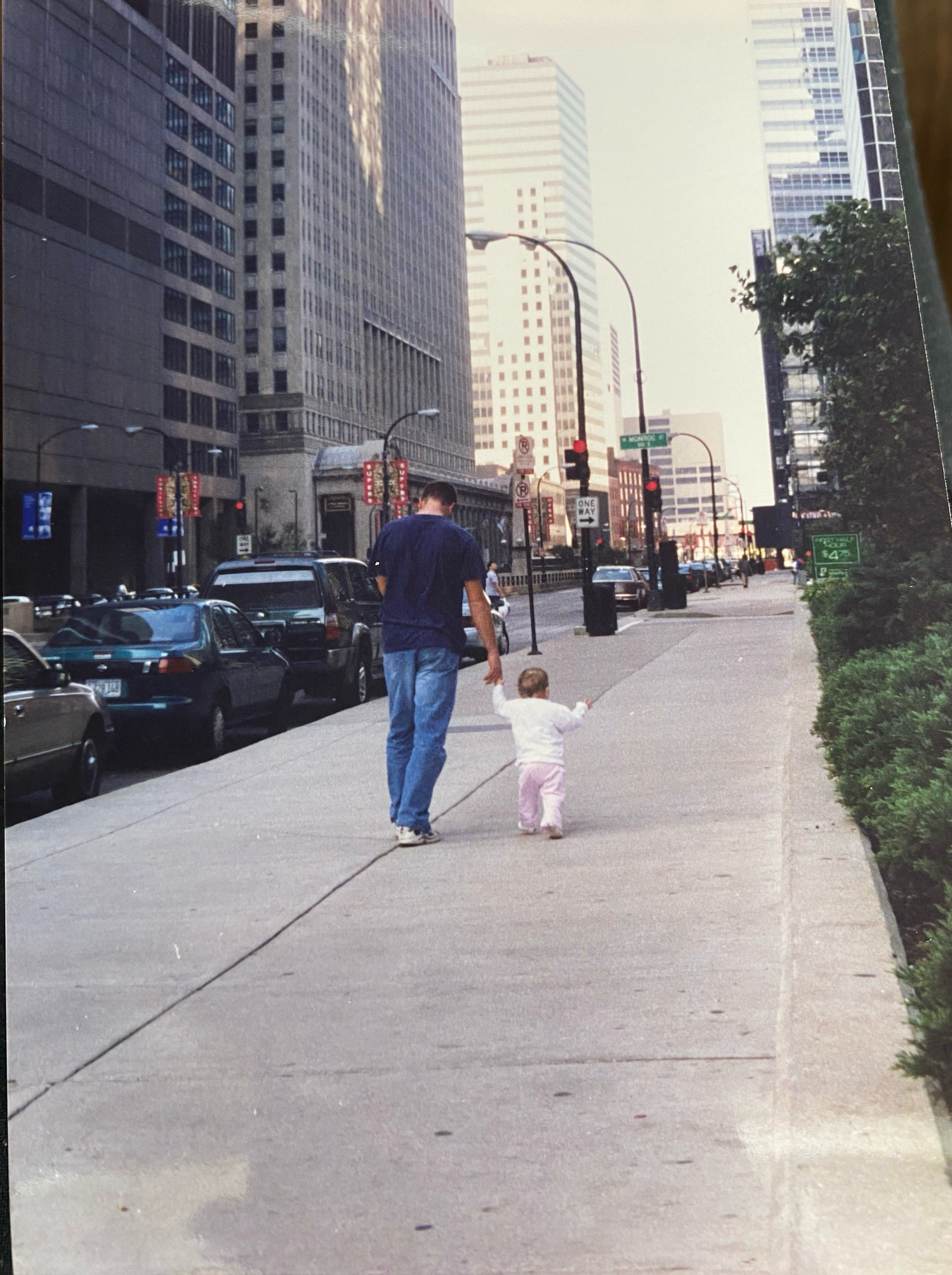 Pair walking together down an urban sidewalk past tall office buildings and parked cars on a city street.