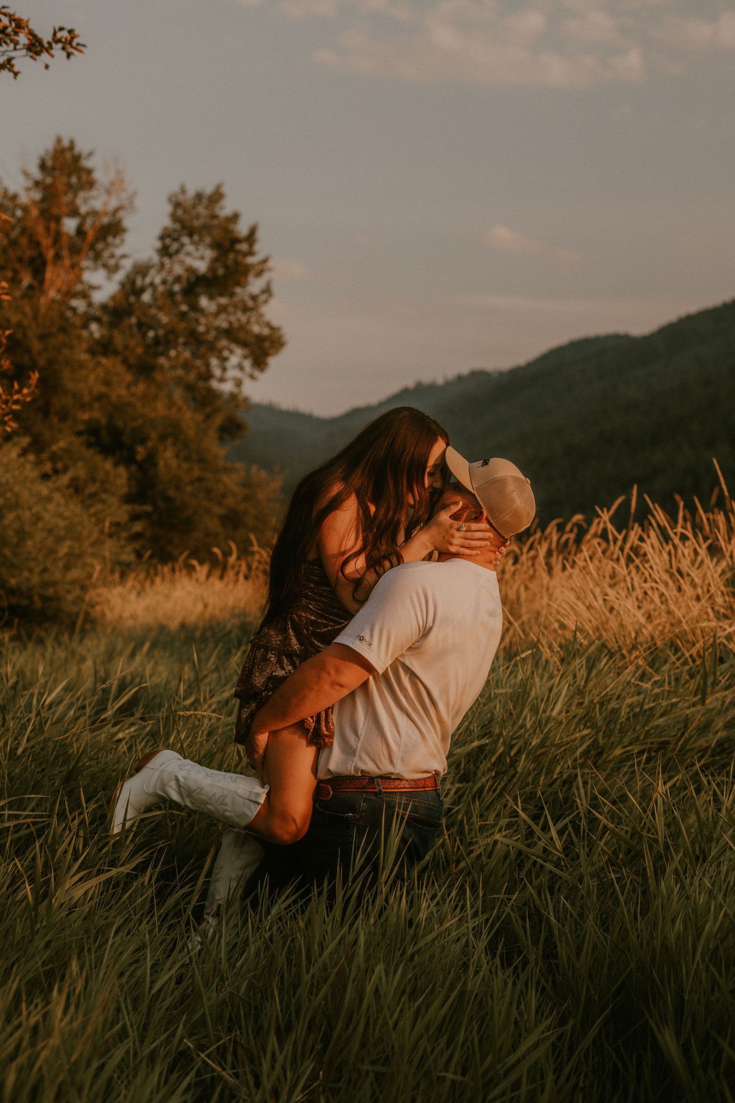 A couple shares a romantic kiss in a golden sunlit meadow with mountains and trees in the background.