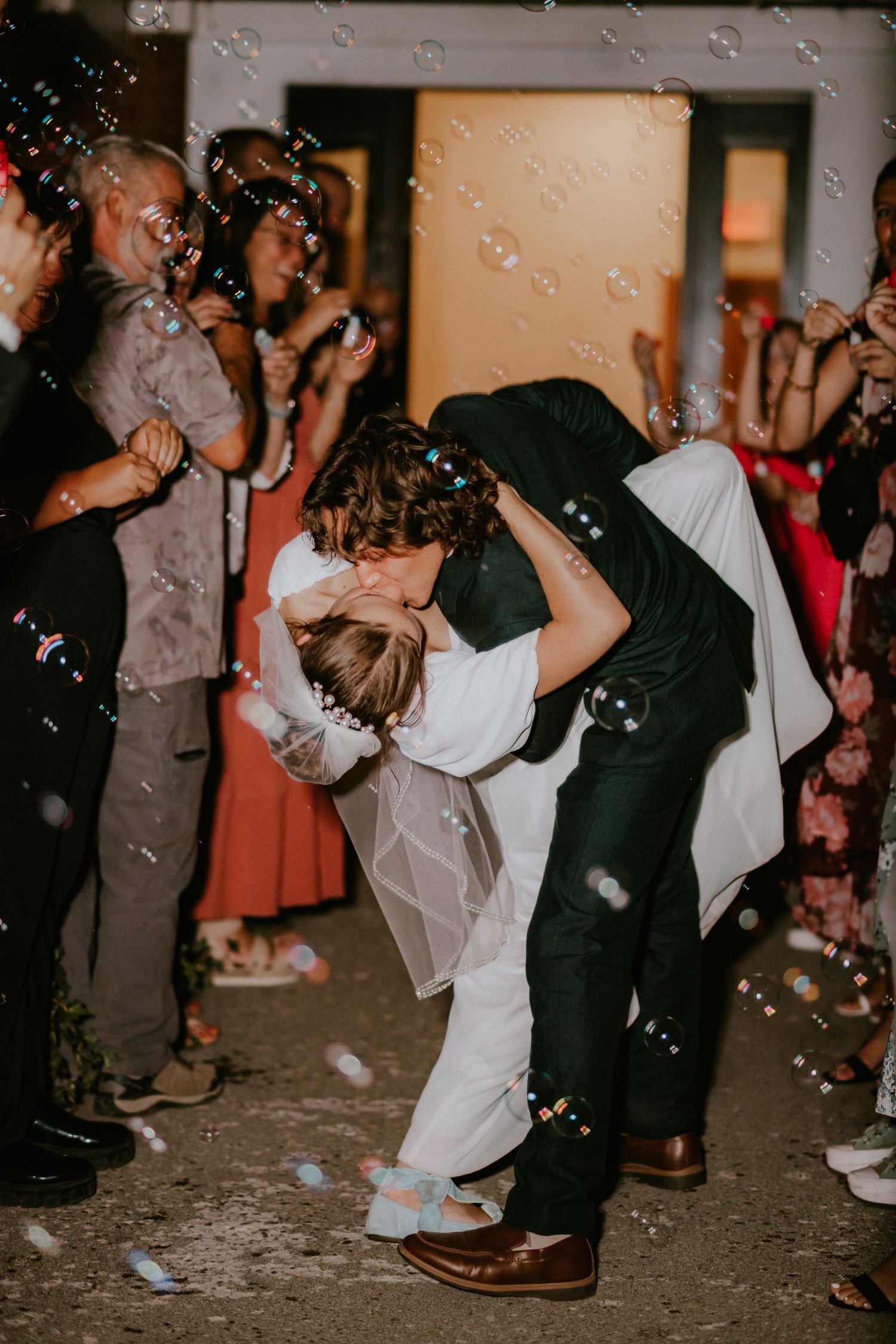 Guests enjoy dancing and dipping on a wedding reception dance floor as bubbles float through the air in moody lighting.