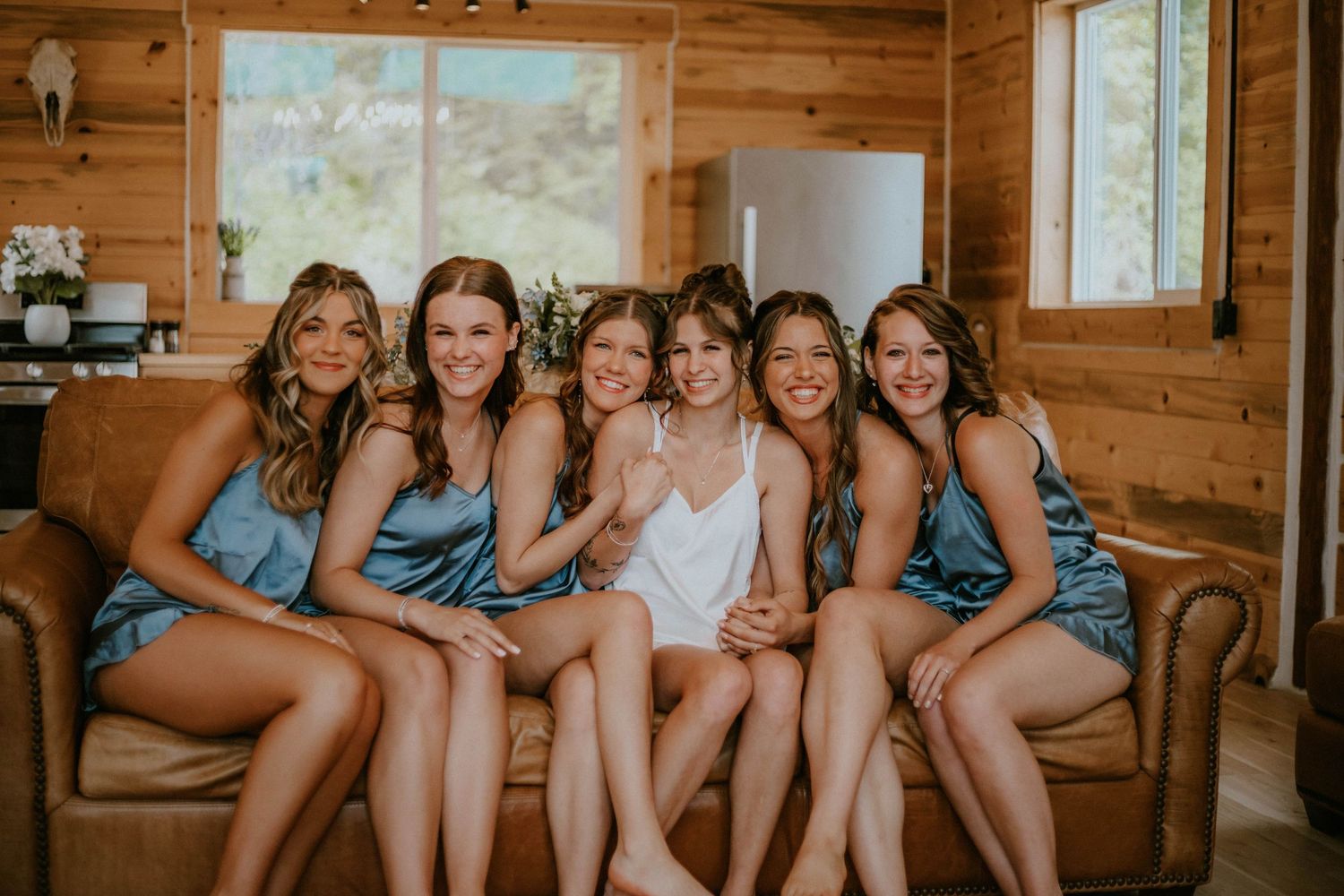 A group of friends in matching blue bridesmaid dresses sit together on a brown leather couch in a rustic cabin setting.