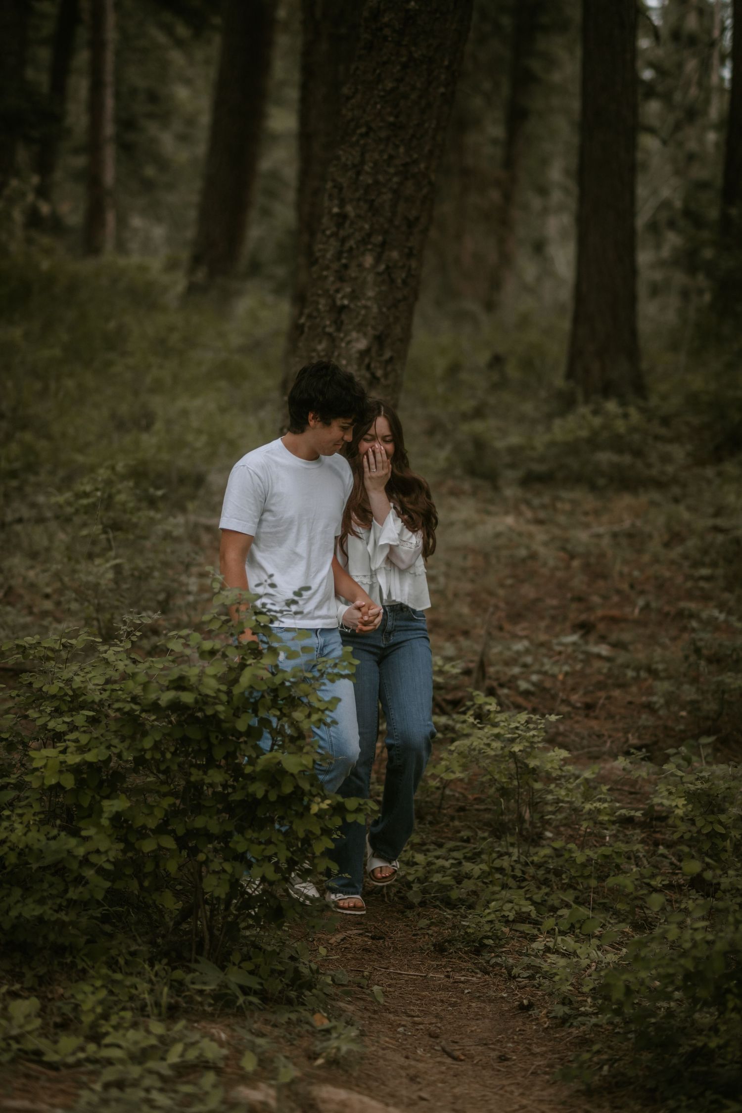 A couple walks hand in hand on a forest trail, smiling and laughing, dressed in white tops and blue jeans.