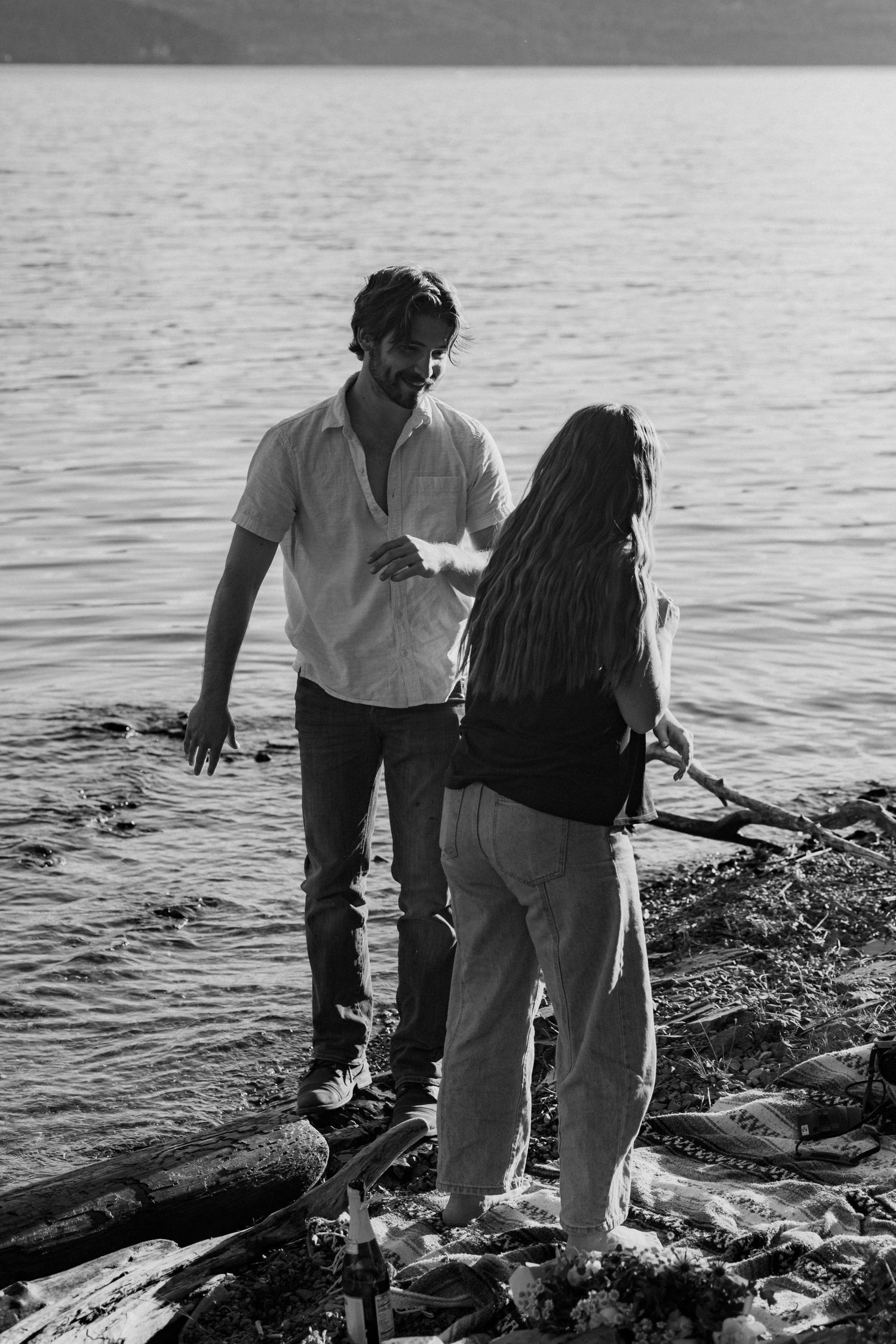 Two people walk along a rocky shoreline near calm water in black and white.