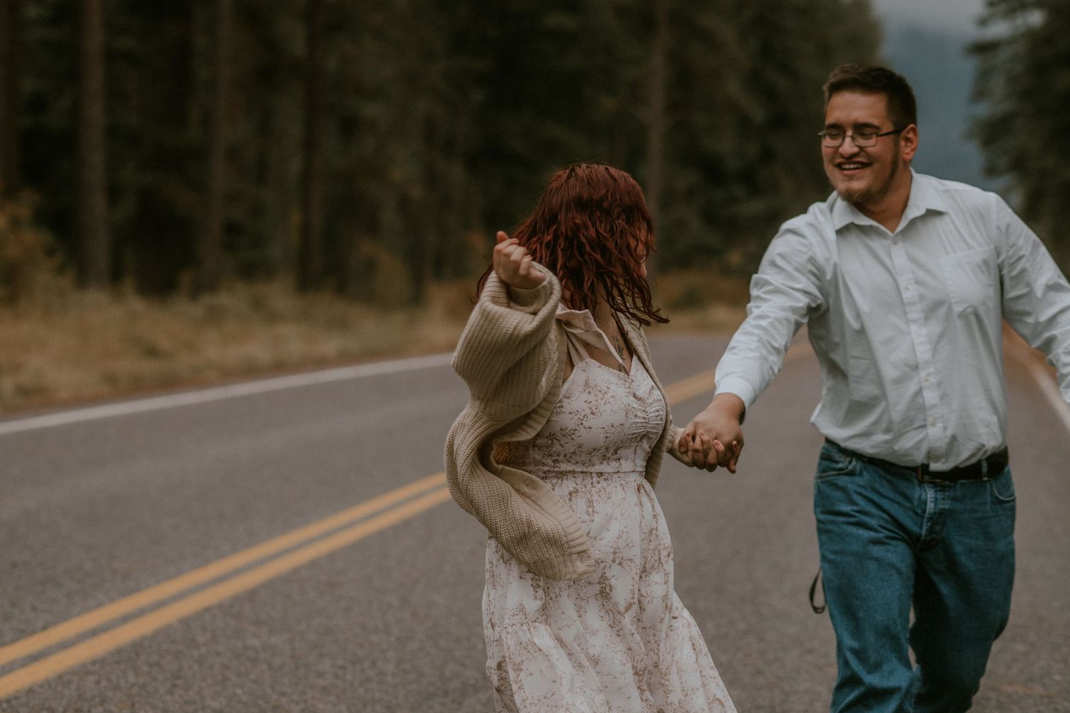 A couple in wedding attire holds hands while walking down a winding forest road surrounded by tall evergreen trees.