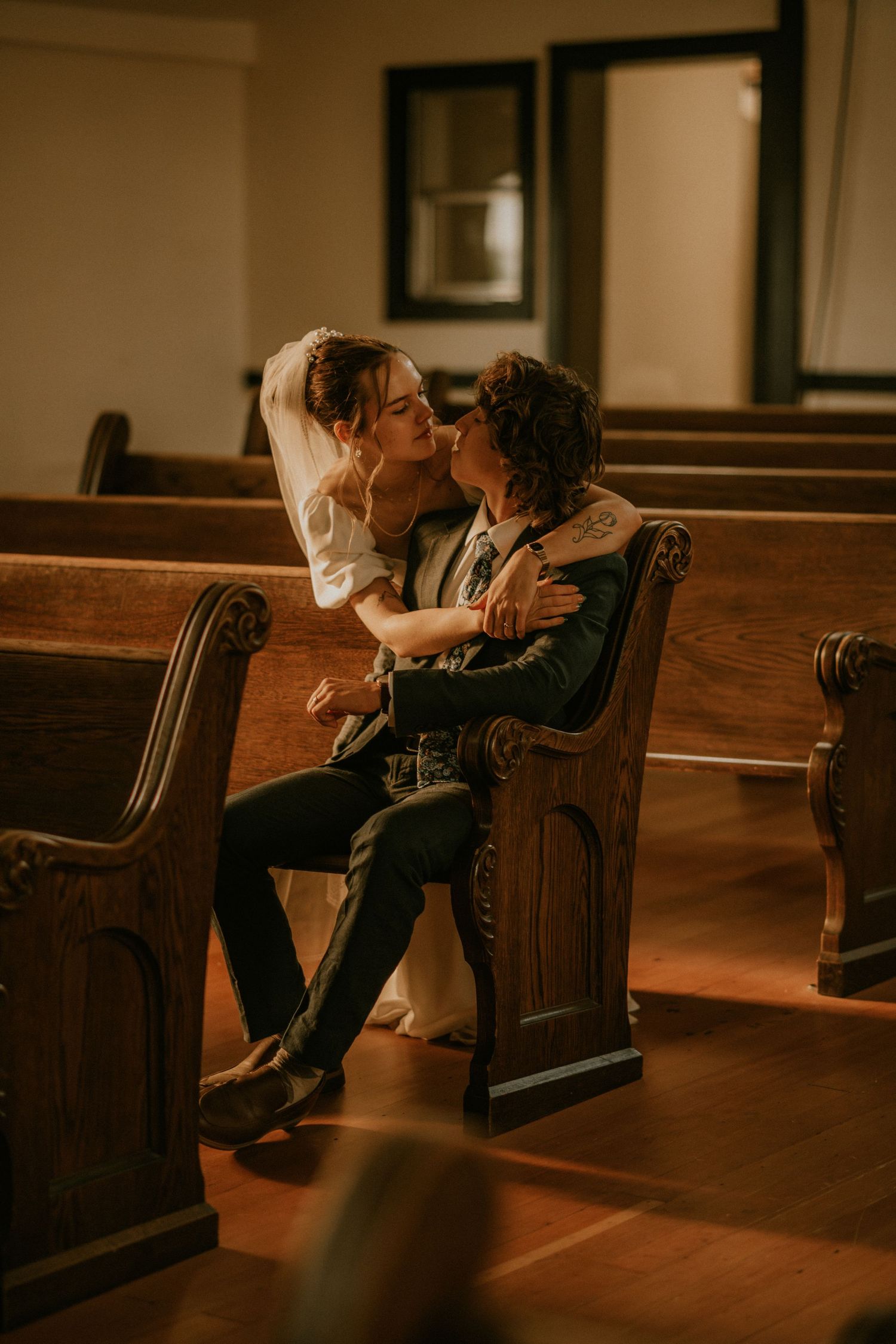 Two people sit together in antique wooden church pews under warm lighting in an intimate setting.
