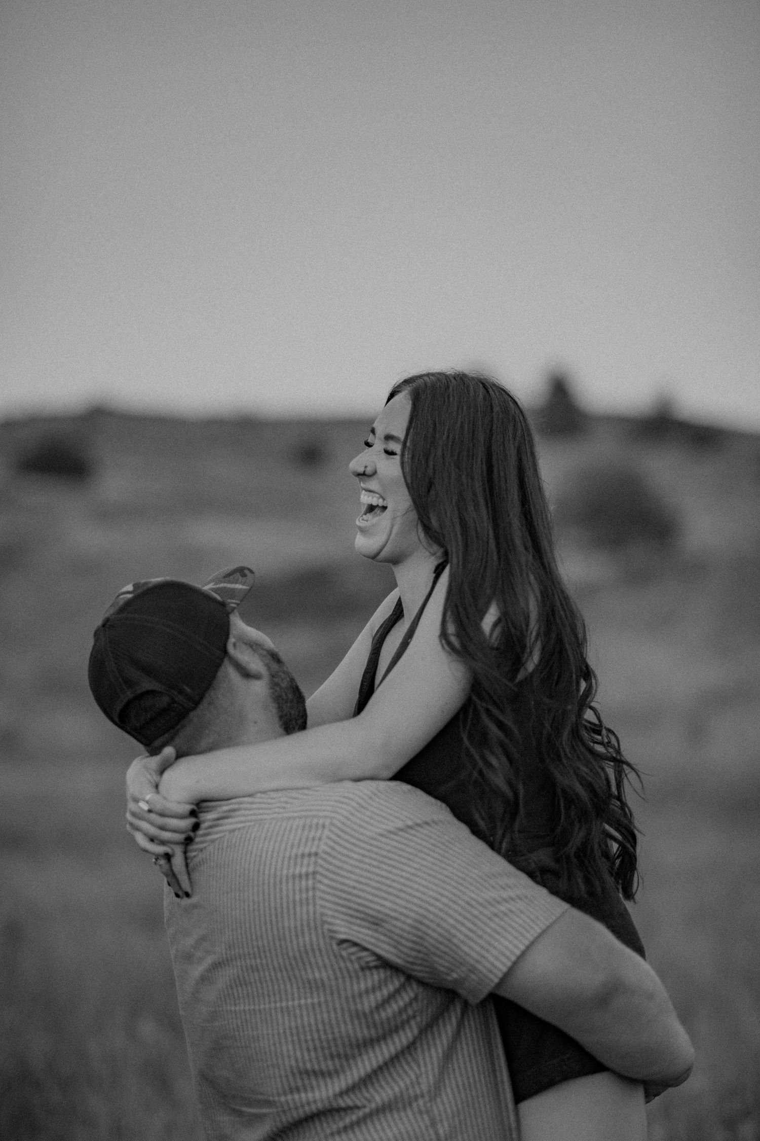 A couple embraces in silhouette against a soft blurred landscape background in black and white.
