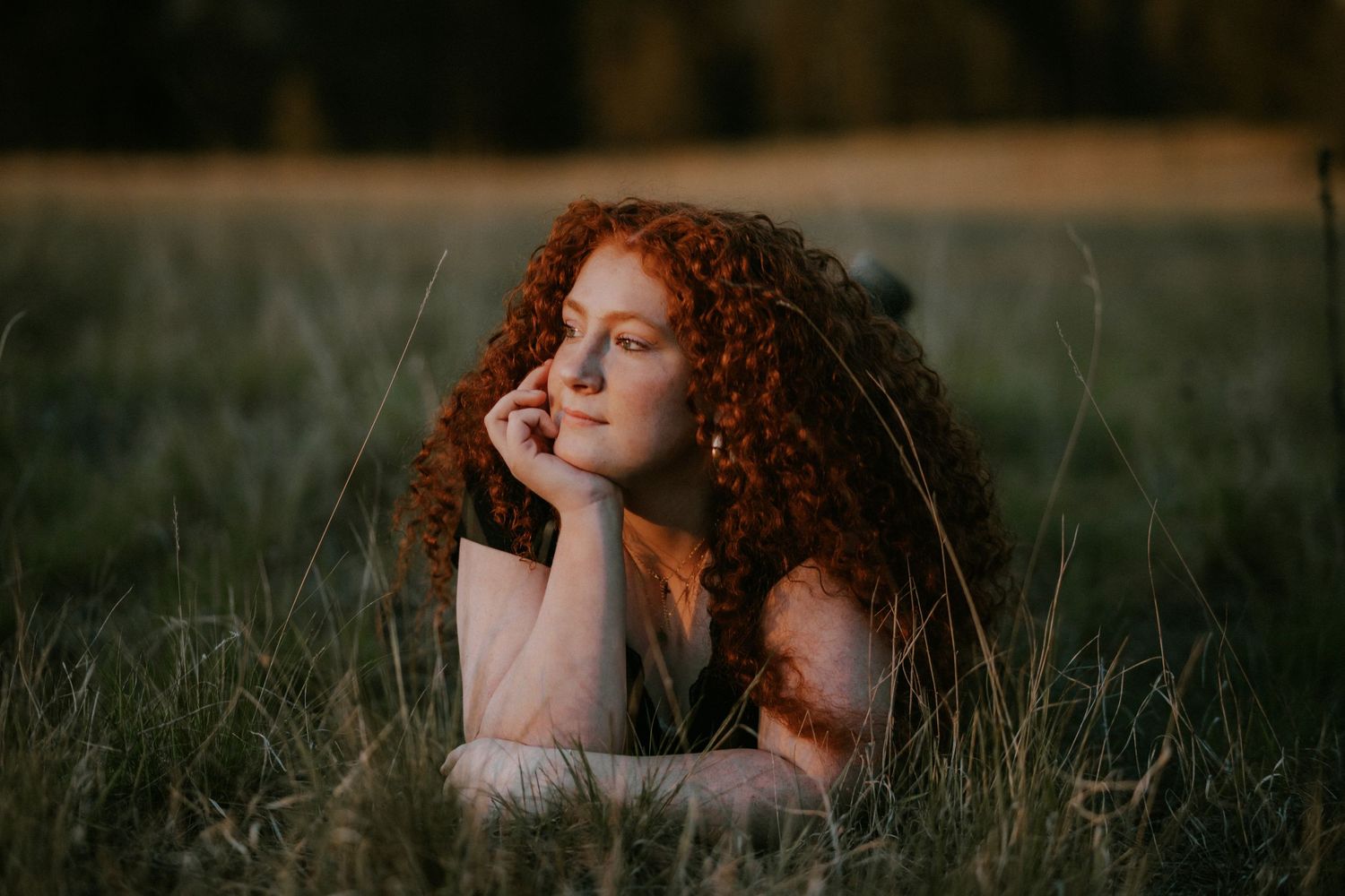 Person with vibrant red curly hair lying peacefully in tall grass during golden hour sunset.