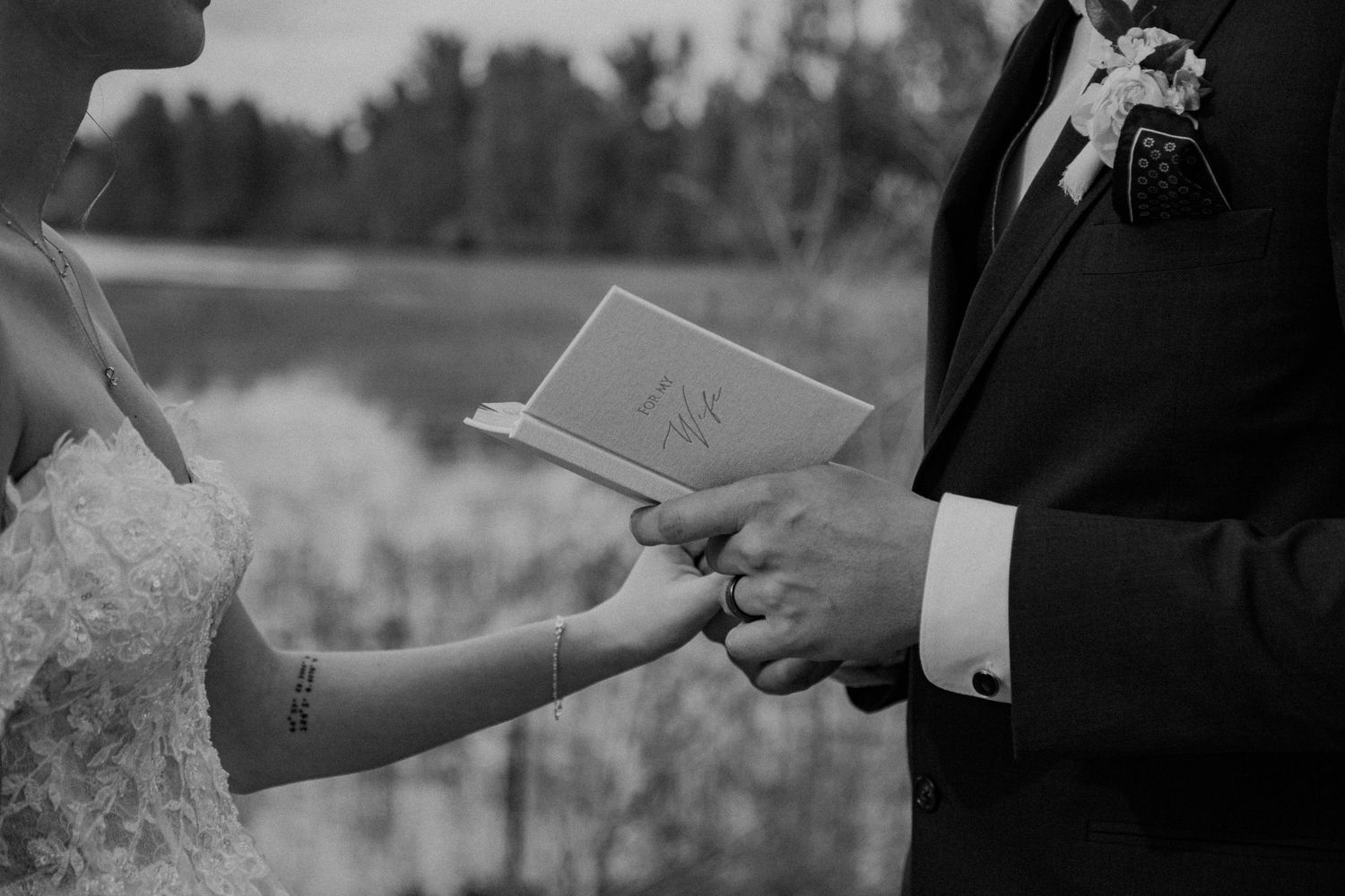 Black and white wedding ceremony photograph showing hands exchanging a note or letter during a lakeside ceremony.