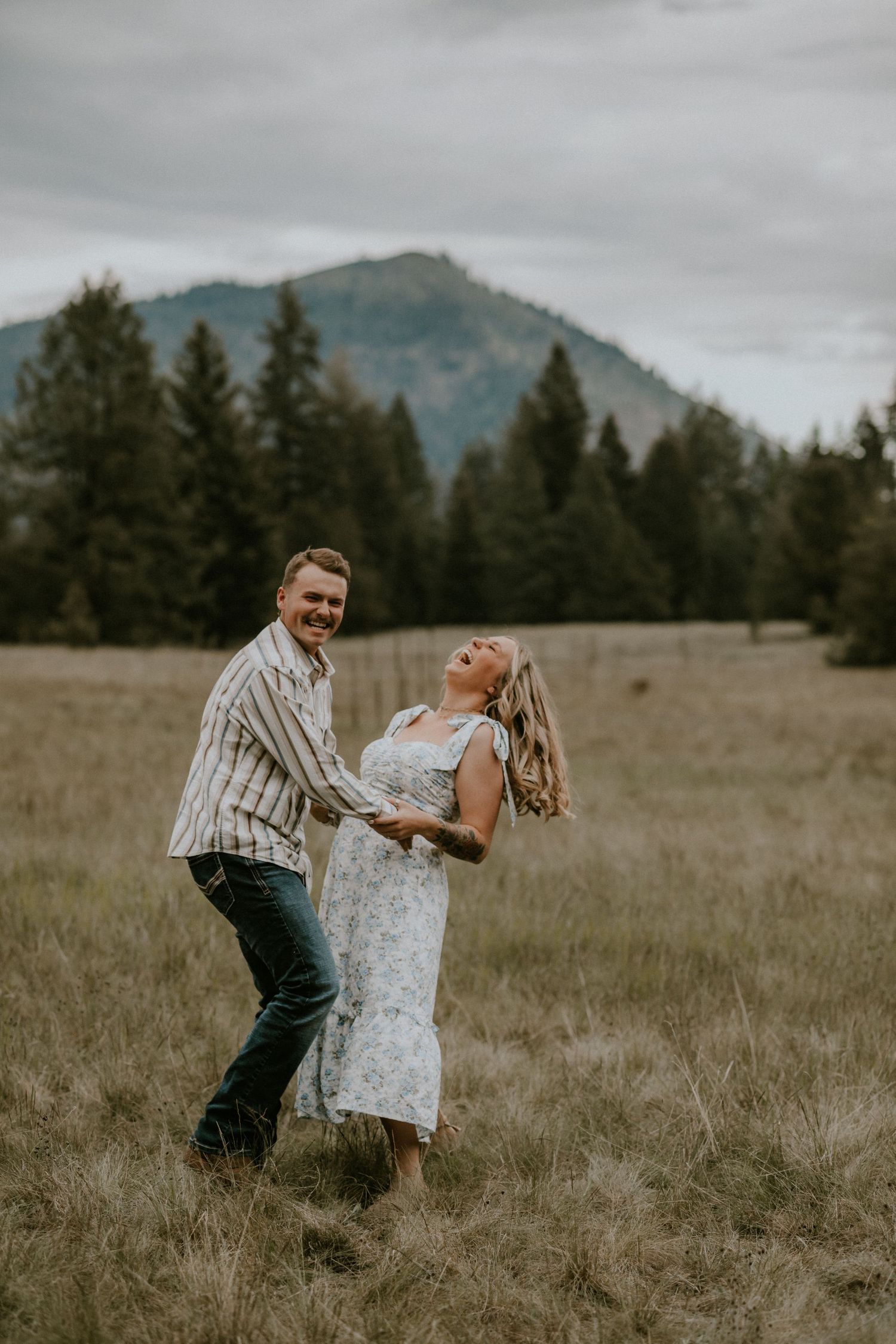 A couple dances and twirls together in a rustic field with mountains and evergreen trees in the background.