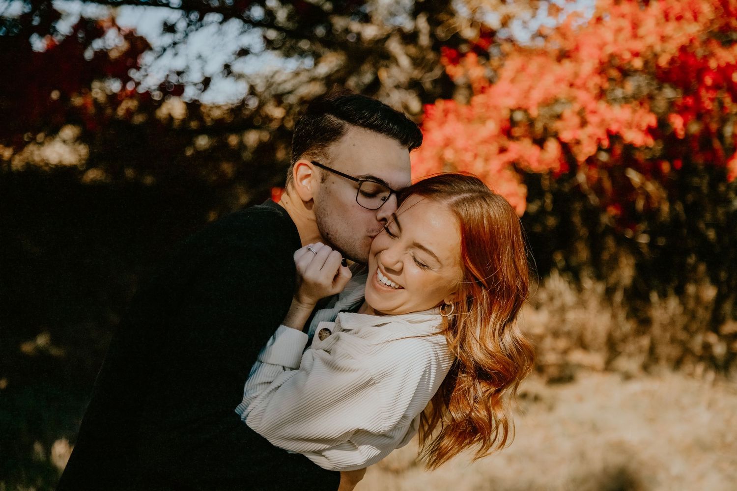 A couple embraces outdoors during autumn with red and orange foliage in the background.