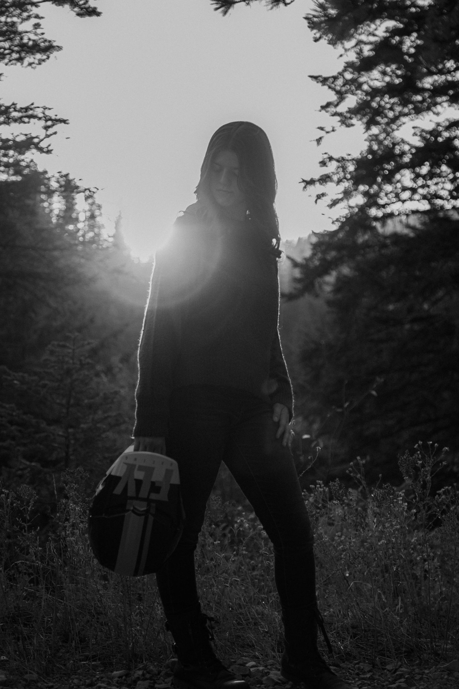 Black and white photo of a person standing in a forest with sunlight streaming through trees behind them.