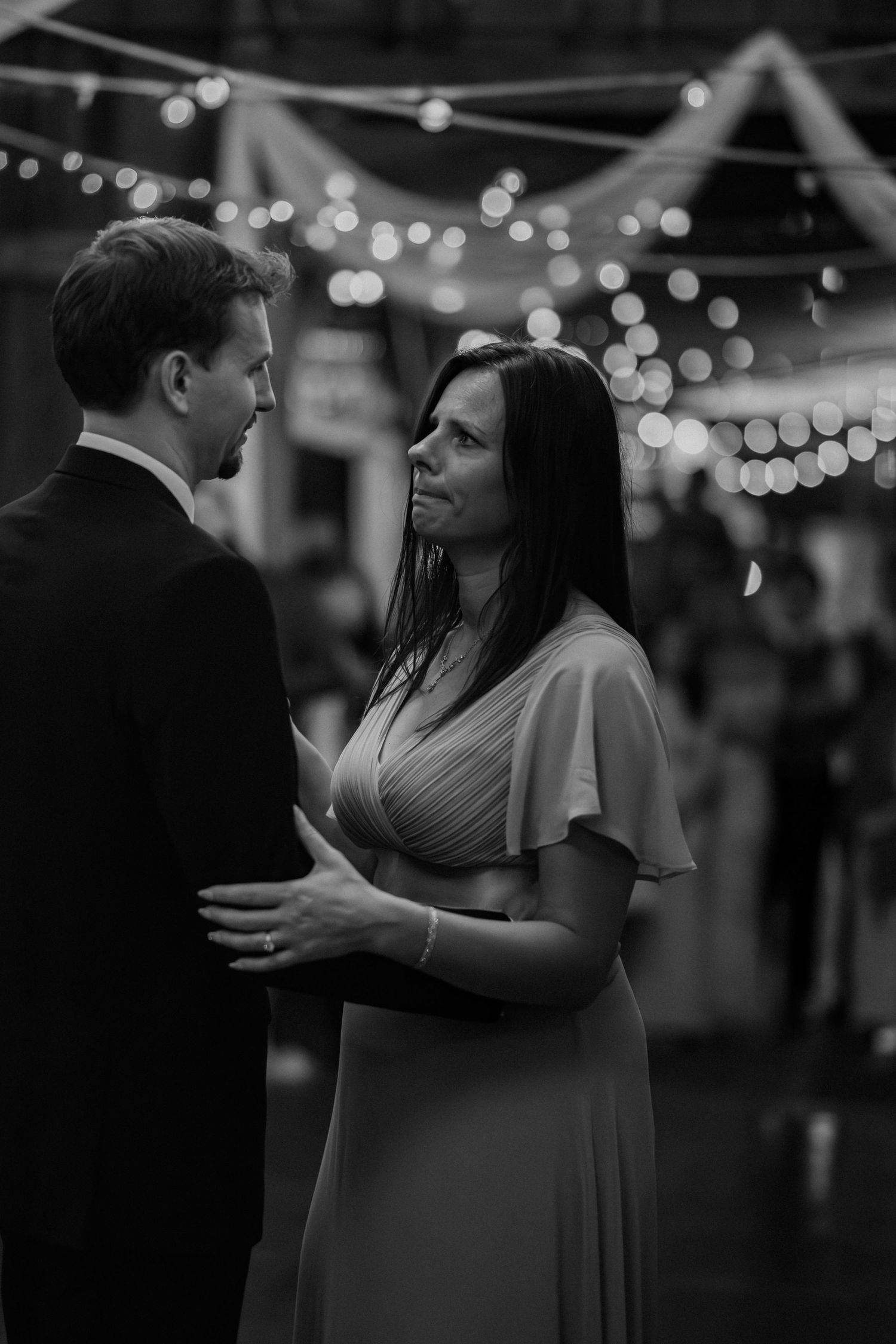 A mom & son shares an emotional moment under twinkling string lights at a nighttime wedding reception.