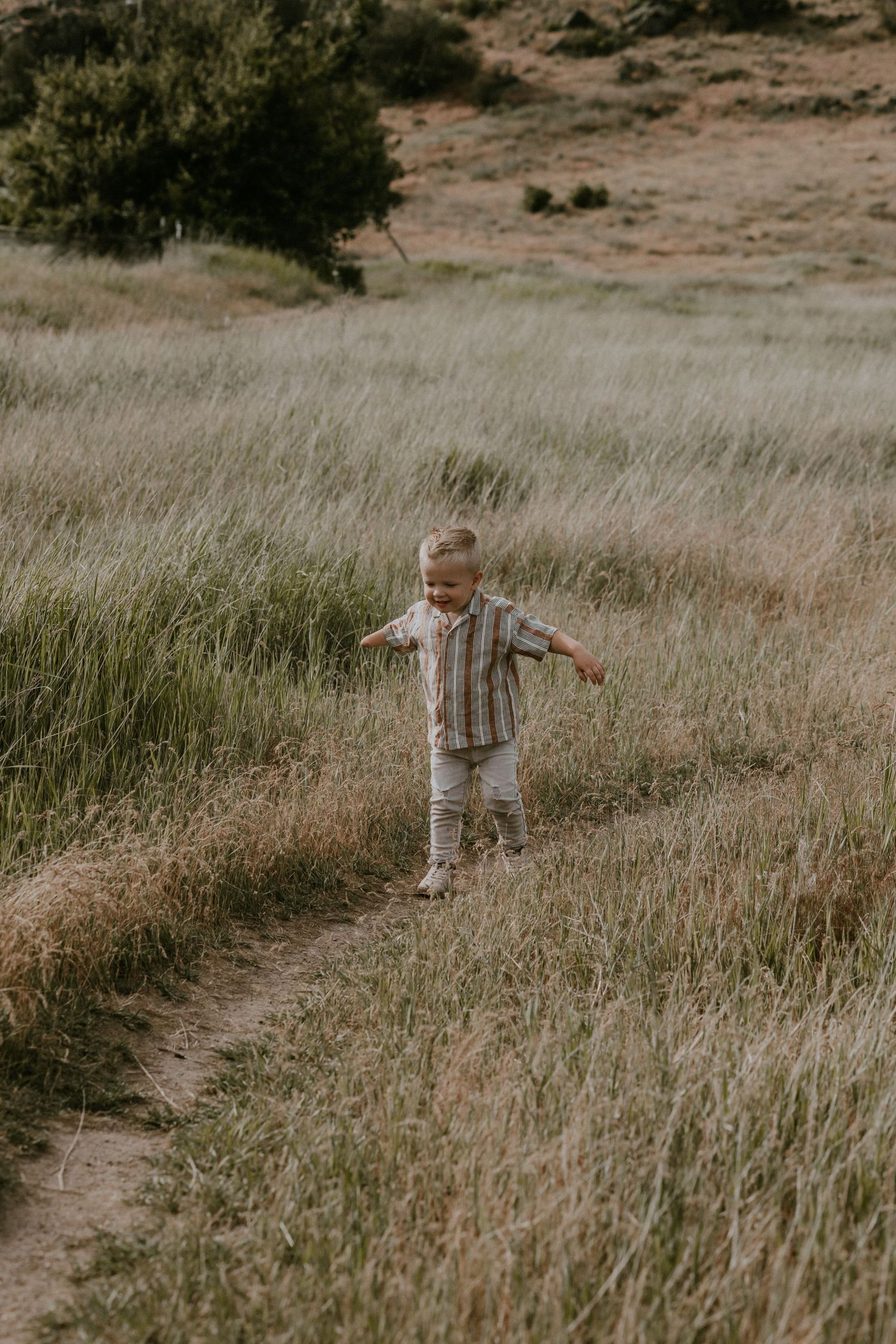 Young child in striped shirt and light pants walks through tall dry grass in a natural outdoor setting.