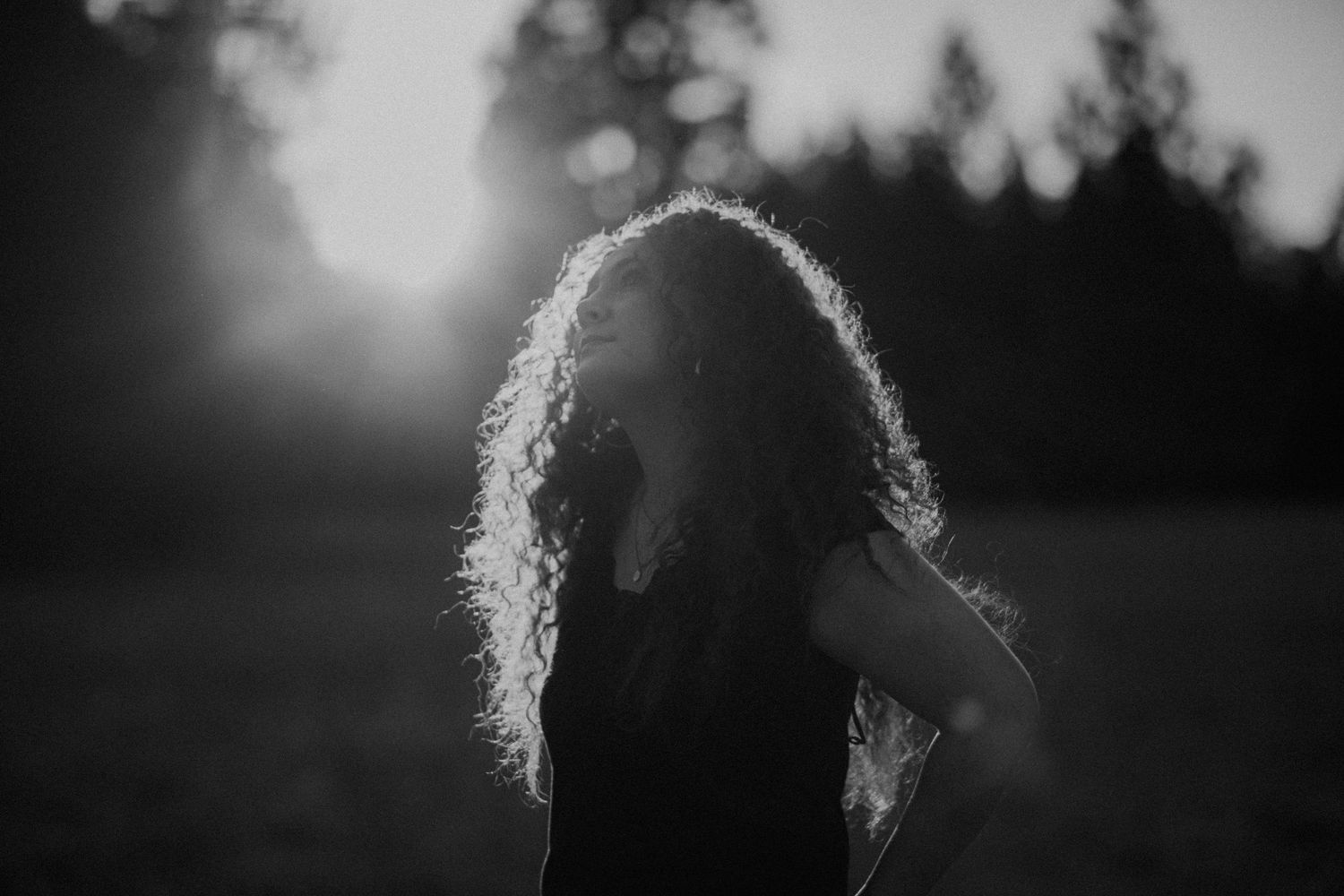 Silhouette of curly-haired figure backlit against sunset with trees in background in black and white photography.