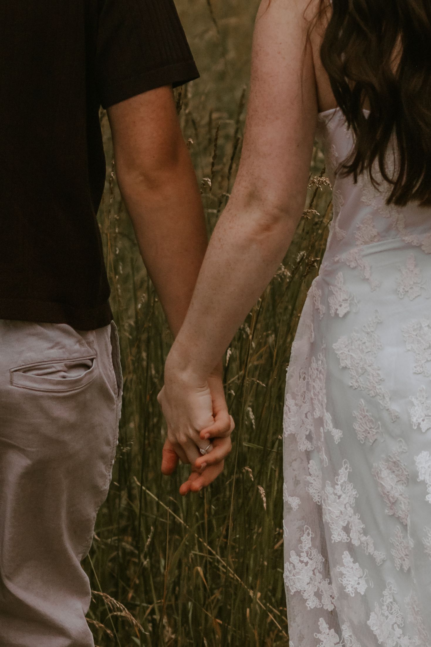 A couple holds hands in a field, the bride wearing a lace wedding dress and an engagement ring.
