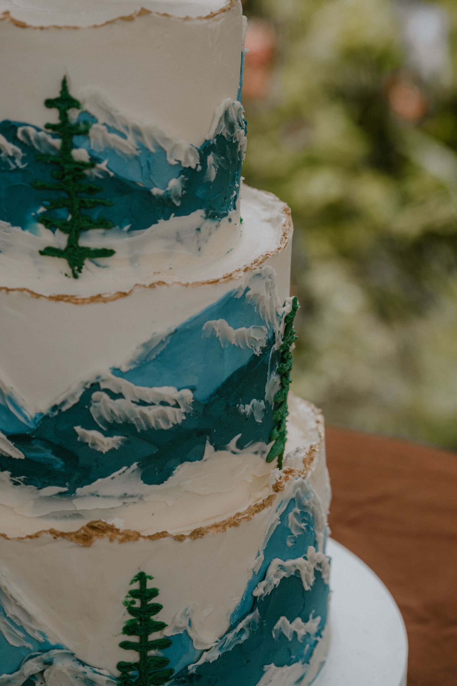 A white wedding cake decorated with teal painted mountains and evergreen trees in an elegant artistic style.