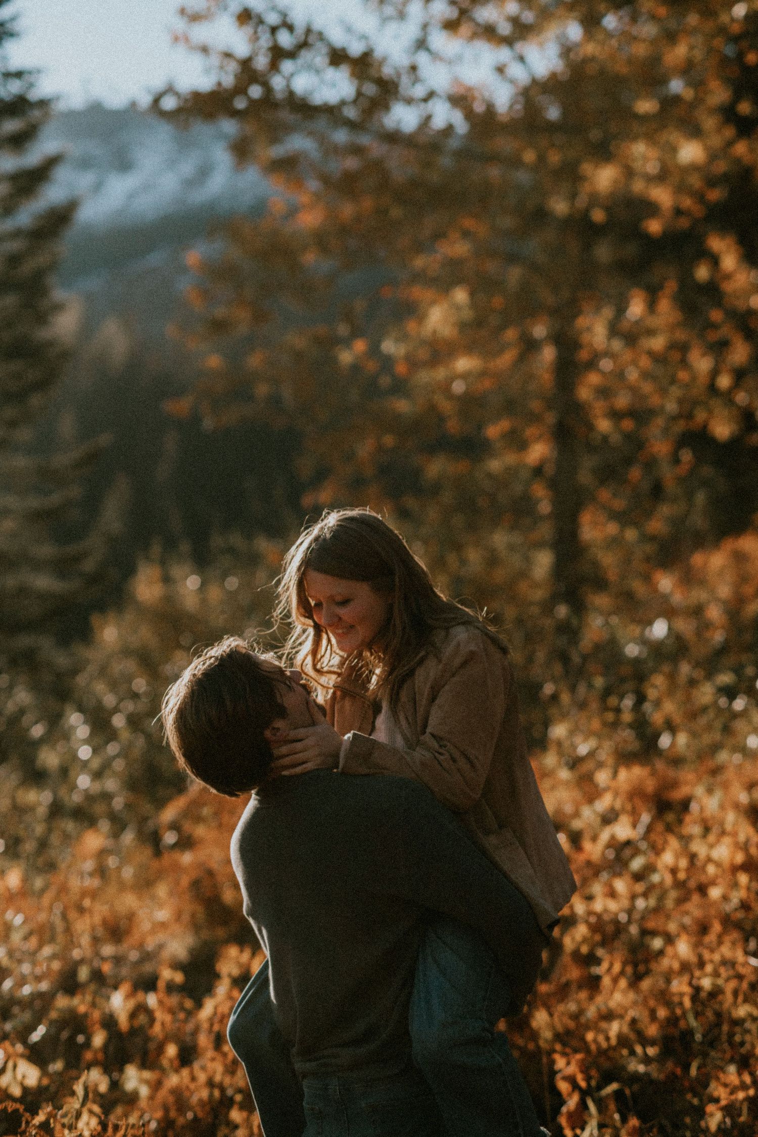 A romantic couple embraces in golden autumn sunlight with fall foliage creating a warm atmospheric backdrop.