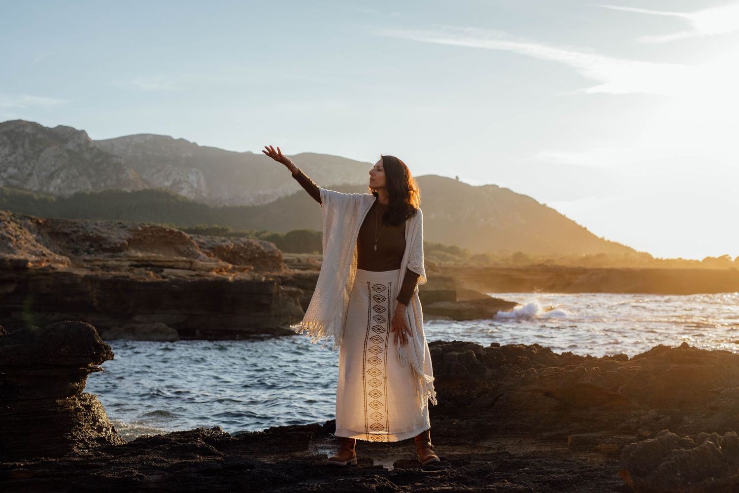 Silhouetted figure in flowing white dress on coastal cliffs at sunset, arms raised in connection. Spiritual photography Mallorca: Soulful