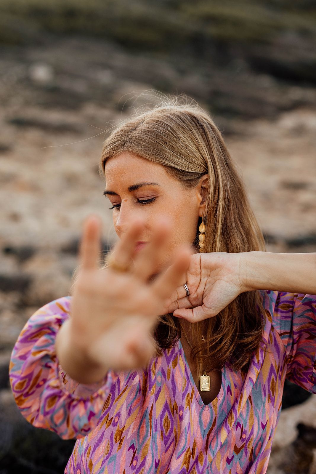 Woman in colorful printed blouse holds hand up to camera while posing against rocky background outdoors.