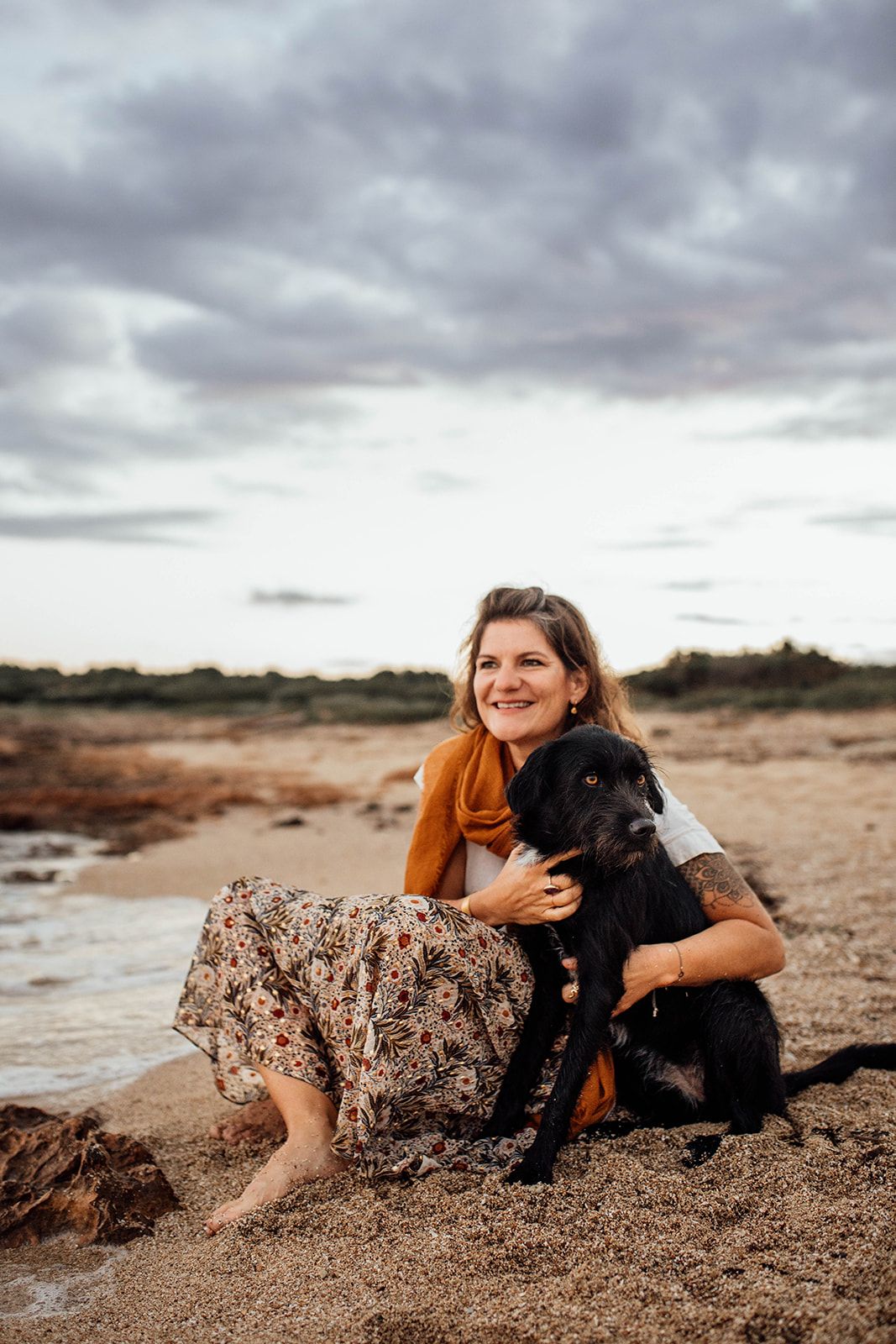 A person in an orange sweater and floral skirt sits on a beach in Mallorca with their black dog under dramatic cloudy skies for a photoshoot