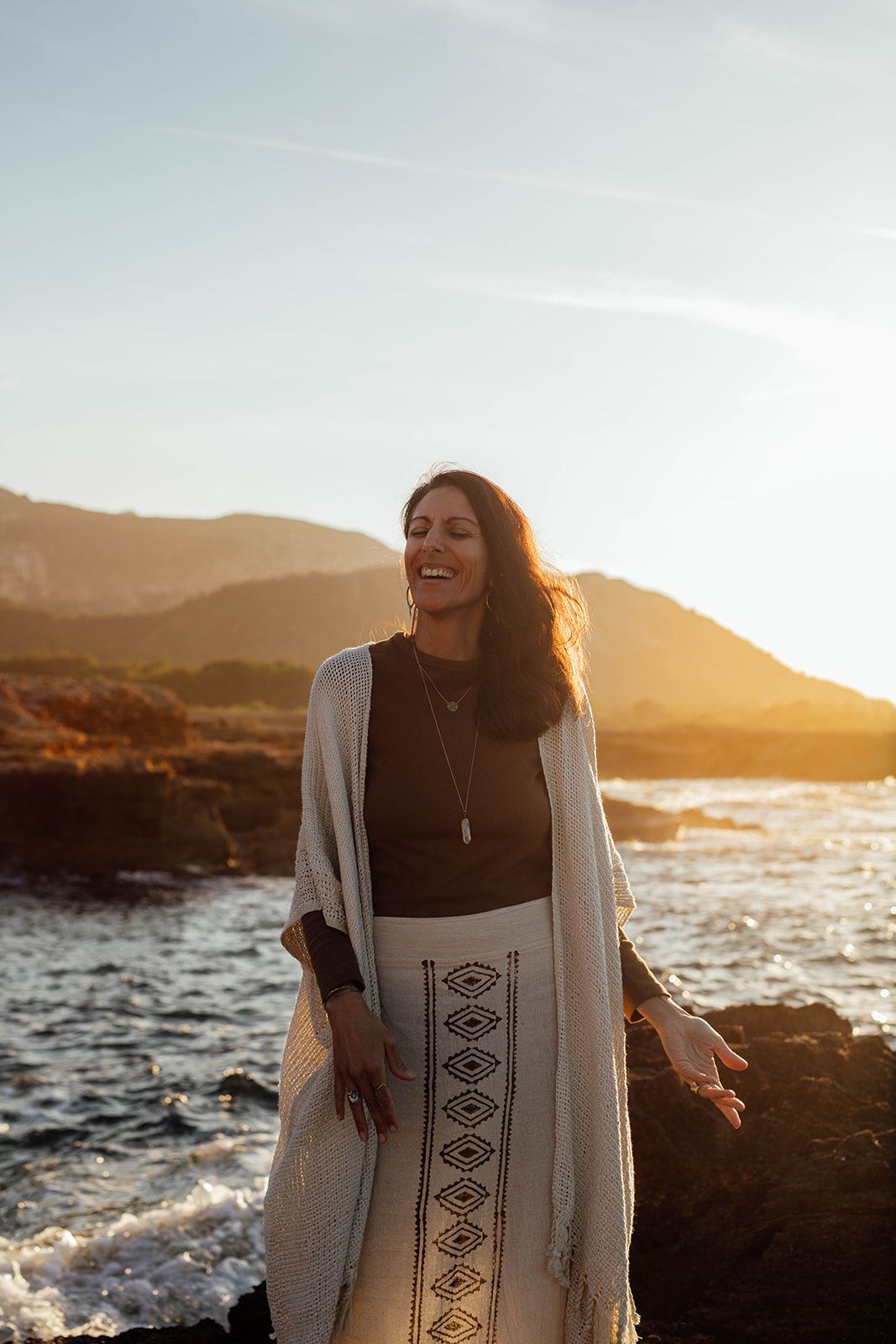 Woman in flowing dress and cardigan smiling by rocky coastline in Mallorca during golden sunset photoshooting.
