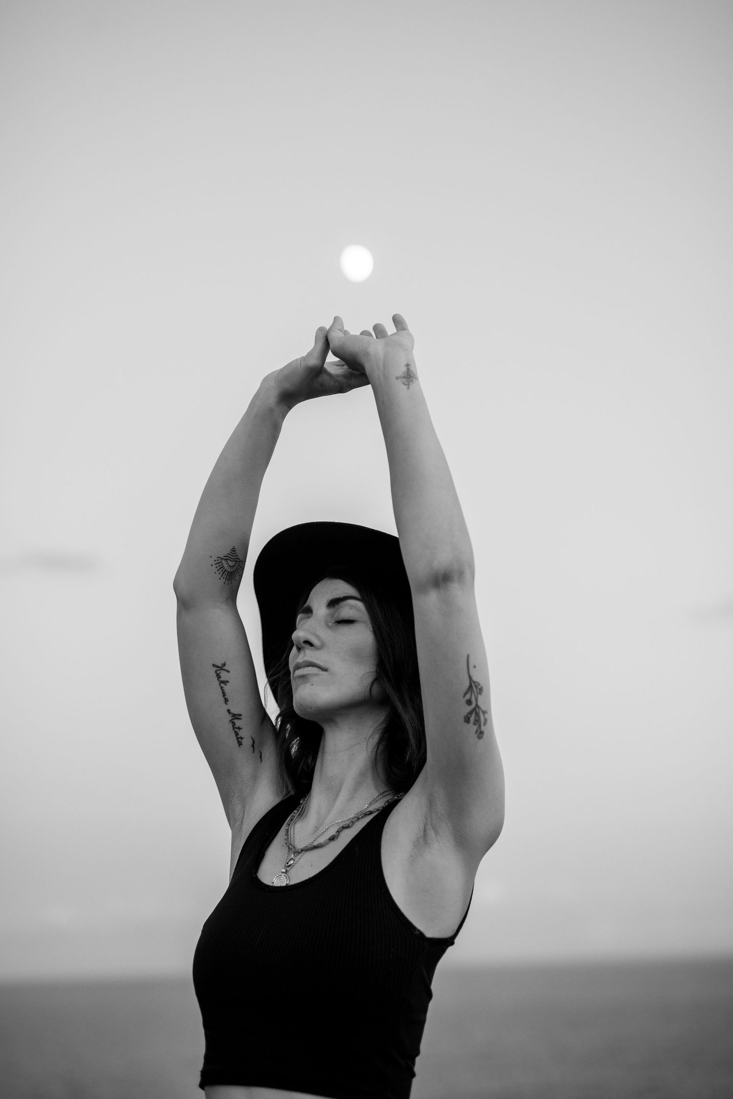 Black and white artistic portrait of person in tank top with raised arms against sky with visible moon in Mallorca.