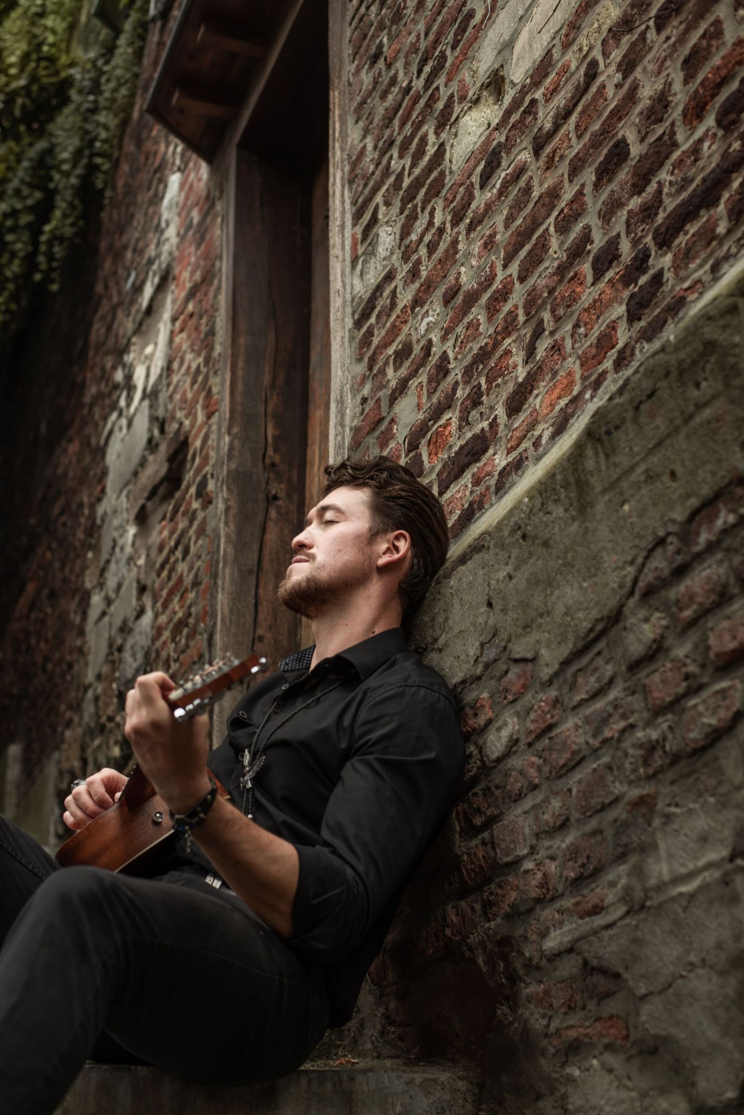 À Liège, portrait d’un homme allongé au sol, dos contre un mur de briques, jouant de la guitare lors d’une séance photo urbaine