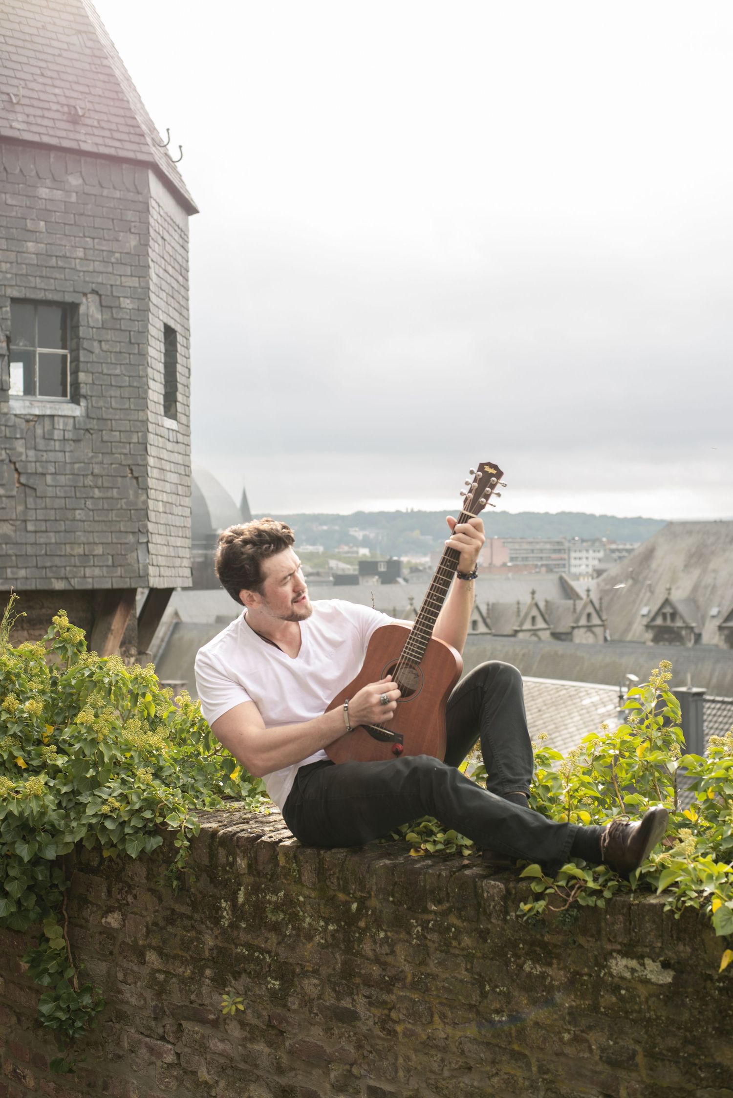 À Liège, sur les hauteurs de la citadelle, portrait d’un homme assis sur un mur avec une vue surplombant la ville, jouant de la guitare lors