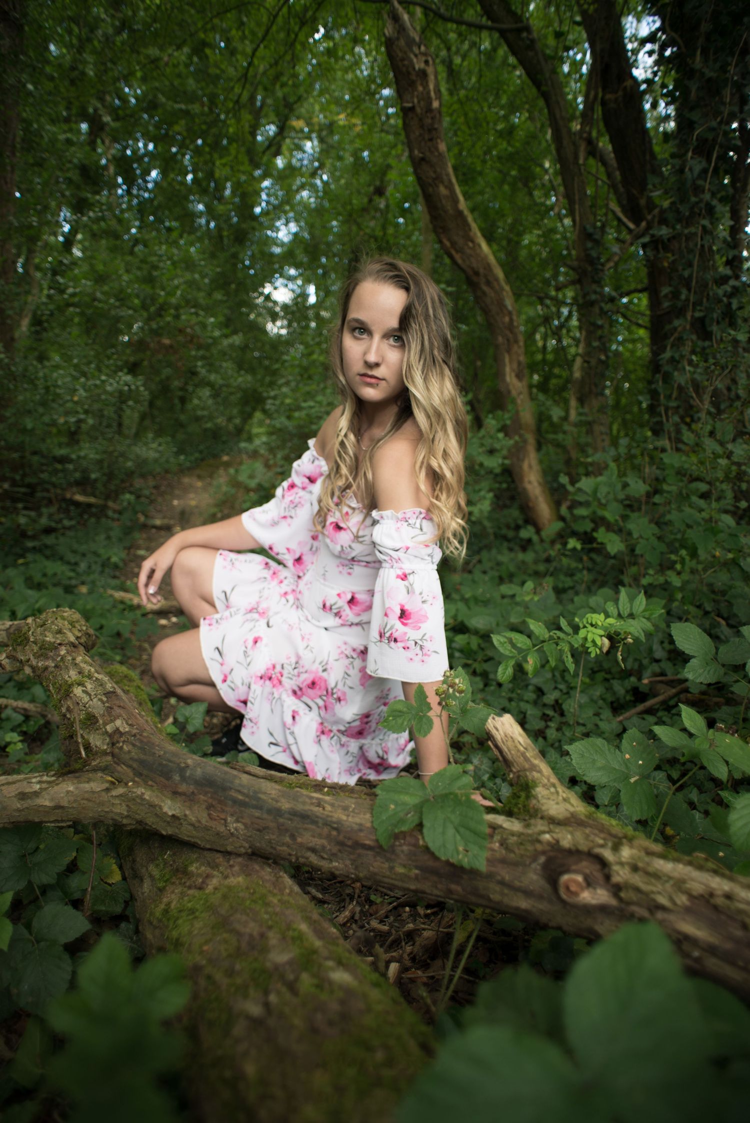 À Ohey, portrait d’une femme accroupie devant une branche avec un fond de forêt verdoyante, regardant la caméra lors d’une séance photo