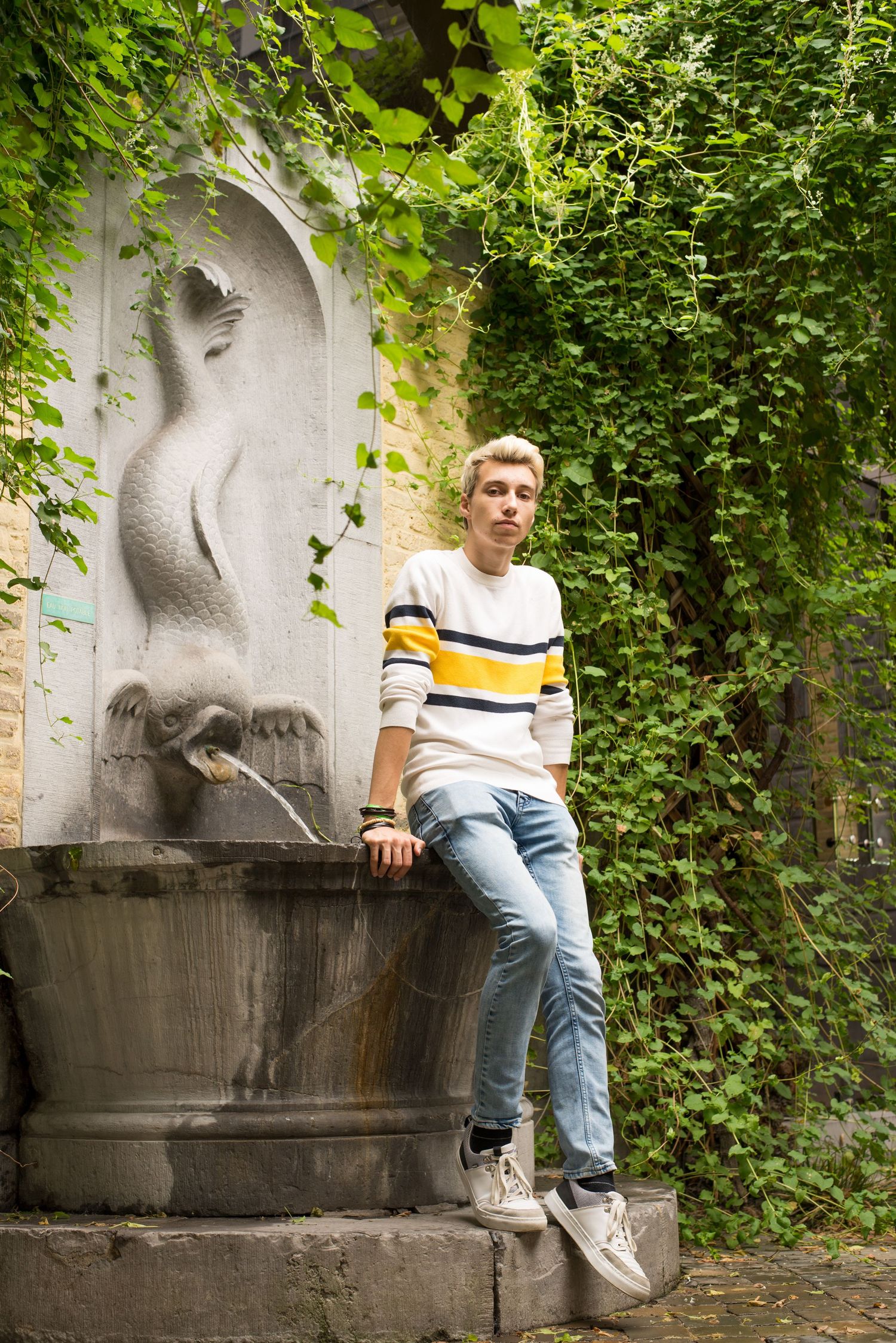 À Liège, dans une cour du quartier Hors-Château, portrait d’un homme assis sur une fontaine entourée de végétation lors d’une séance photo