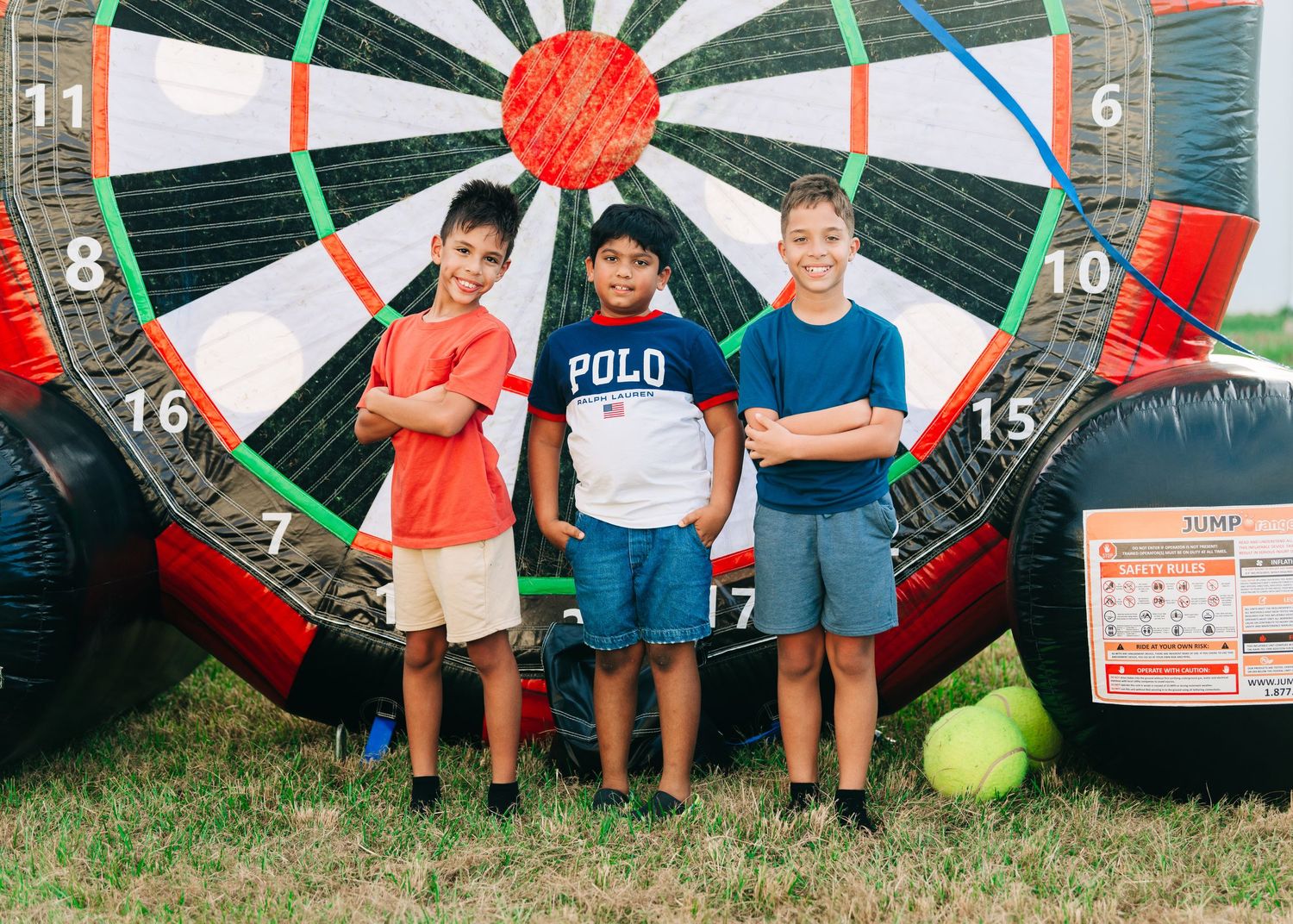 Three boys pose with crossed arms in front of a large inflatable dartboard at an outdoor carnival event.