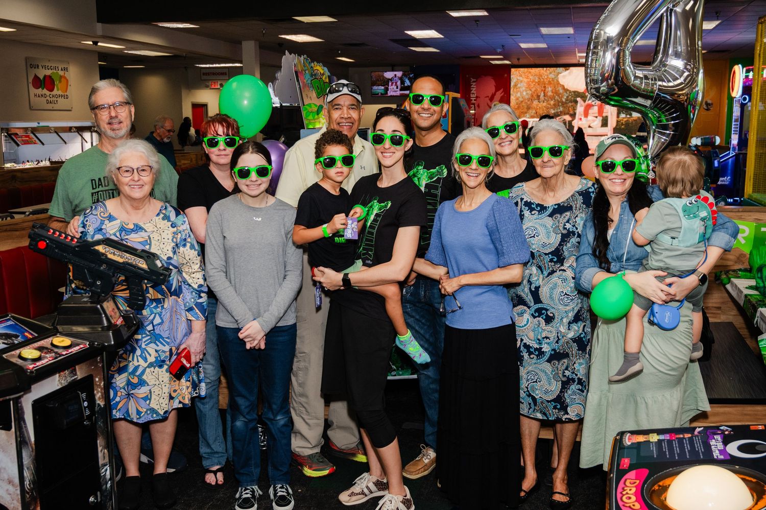 Group of people wearing green sunglasses celebrating at a party with balloons and decorations.