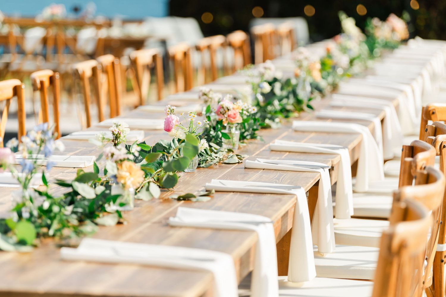 Elegant wedding reception long table with floral greenery runner, pink roses, candles, and wooden chairs in a bright venue.