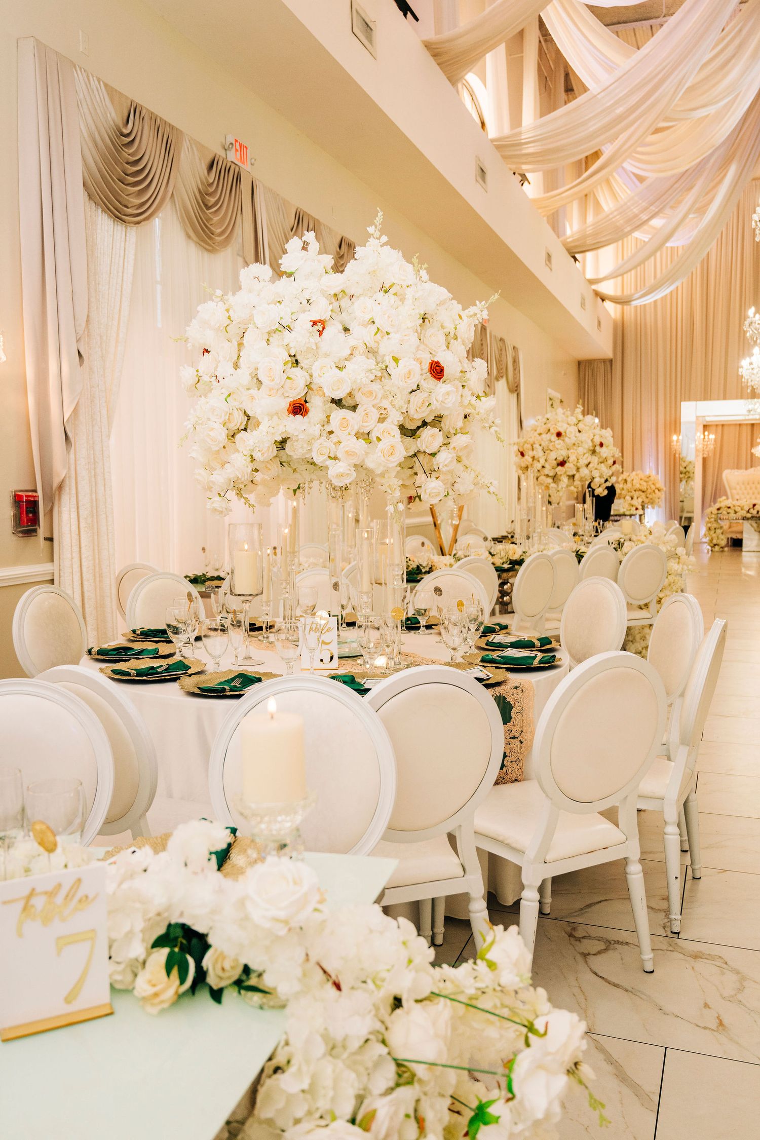 Elegant wedding reception hall with white floral centerpieces, green accents, ivory chairs, and draped ceiling decor.