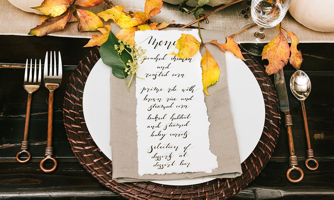 Elegant fall table setting with calligraphy menu, copper cutlery, wicker charger, and autumn leaves decor.