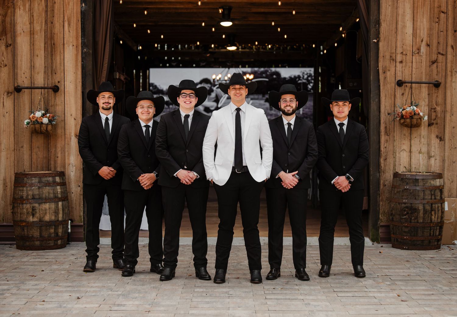Groom in white suit with groomsmen in black suits and cowboy hats posing in rustic barn wedding venue.