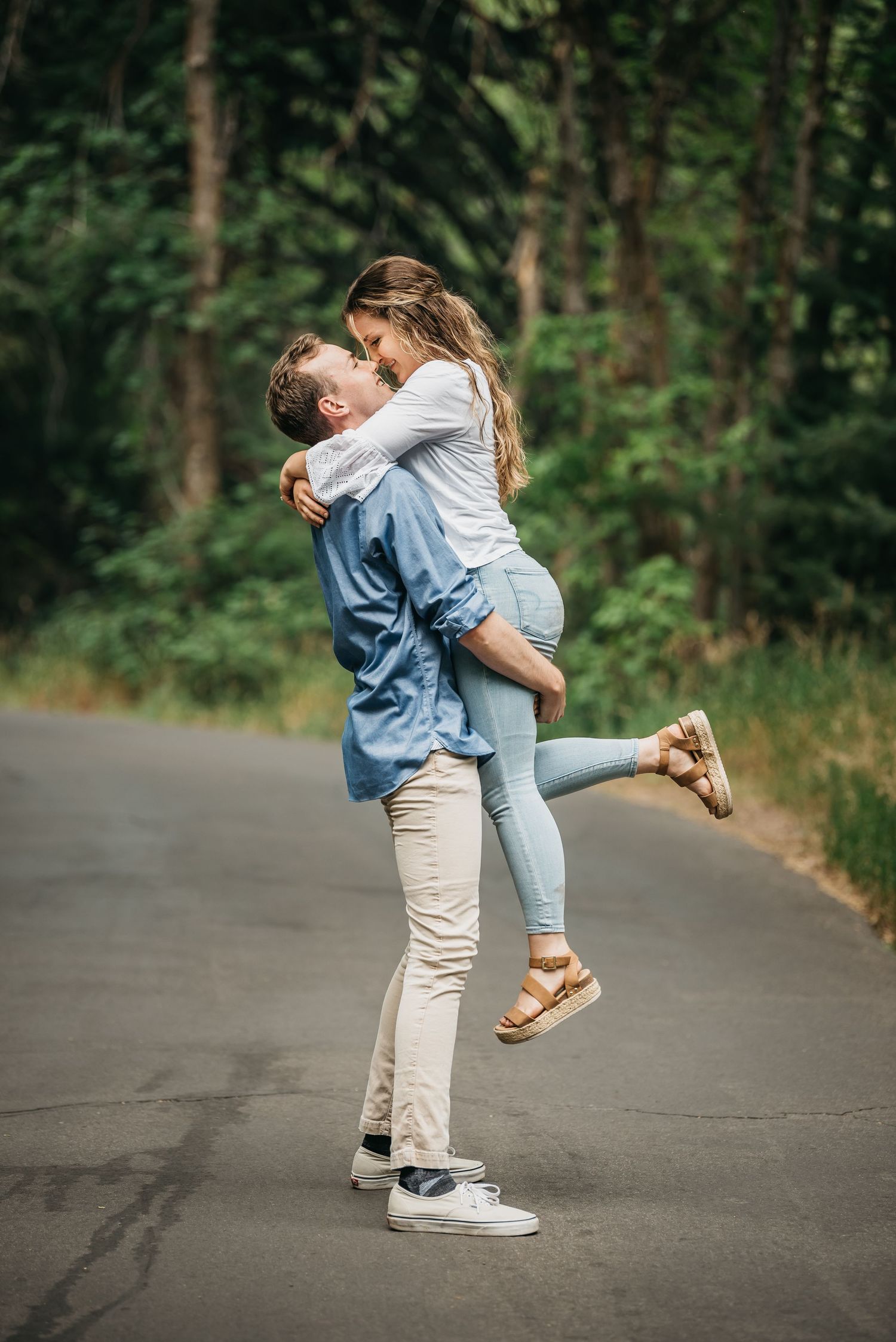 A man lifts a woman in his arms on a road surrounded by green trees, sharing a romantic moment.