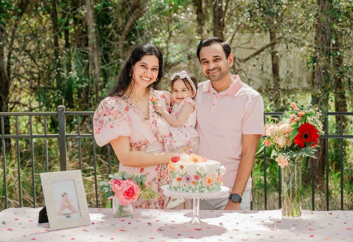 Family of three smiling behind a floral birthday cake outdoors, parents holding toddler girl in pink dress at garden party.