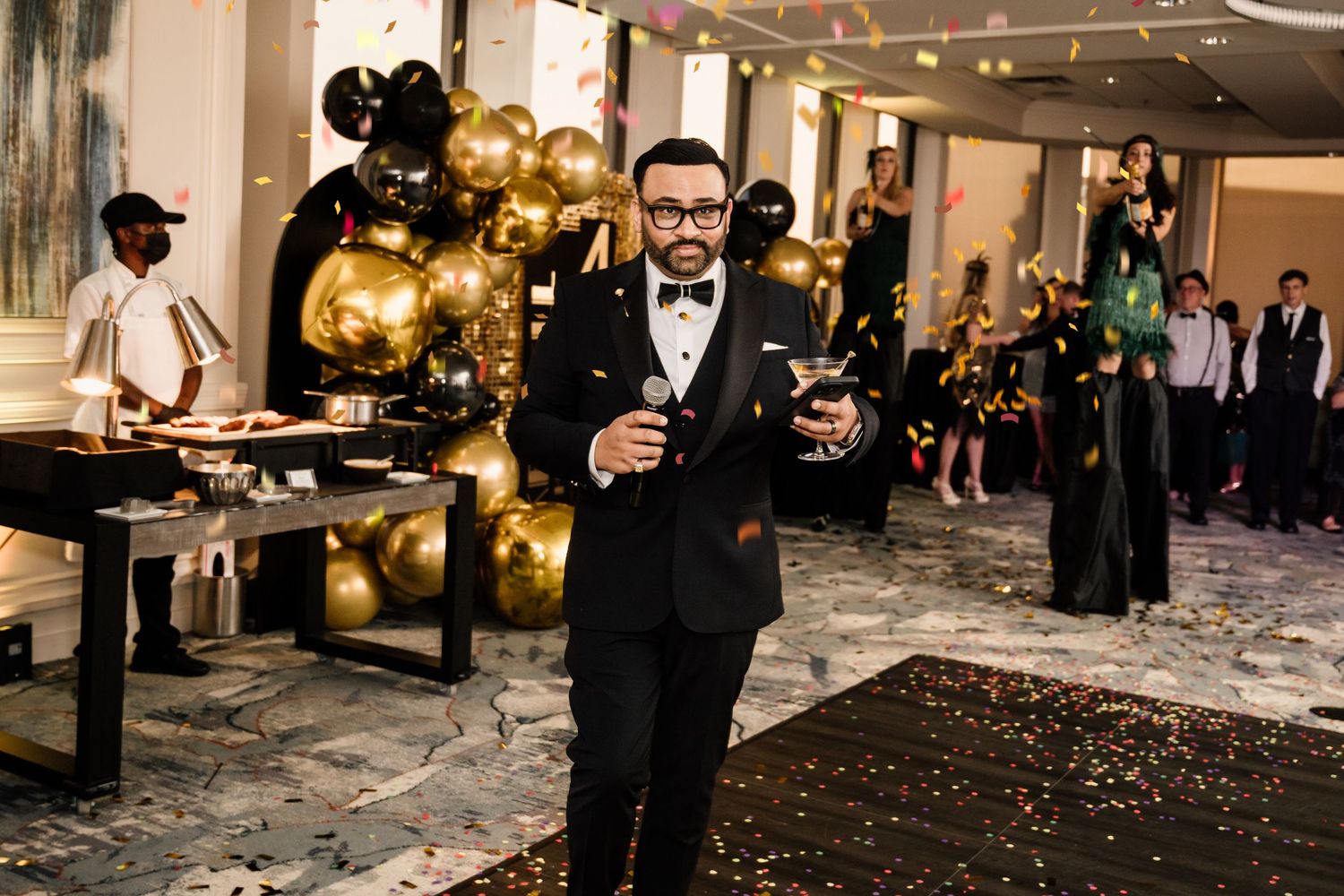 Man in tuxedo holding champagne at elegant New Year's Eve party with gold balloons and confetti.