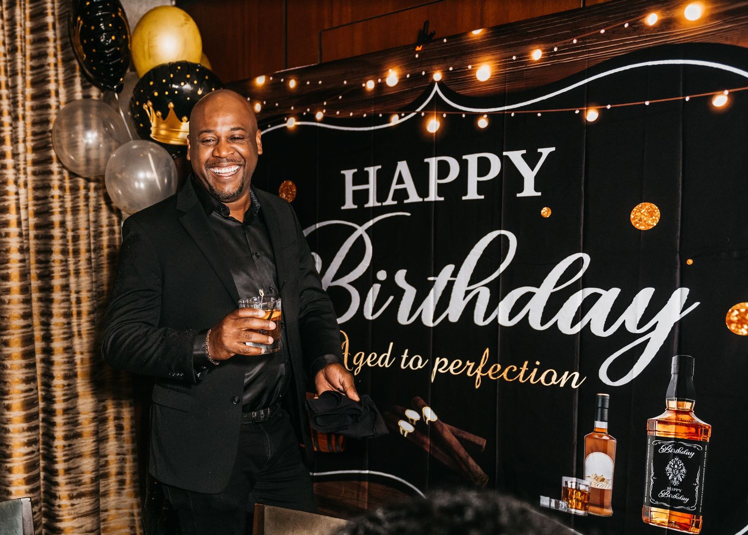 Man in black suit smiling at birthday party with Happy Birthday banner, balloons, and whiskey bottle.