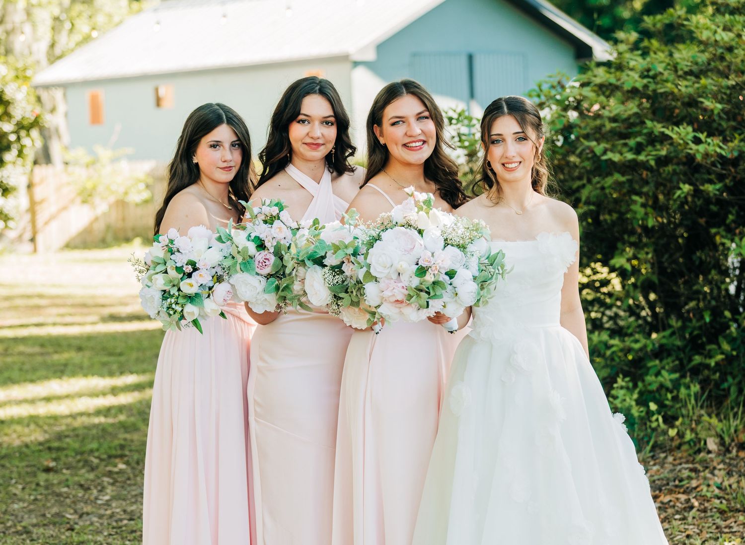 Bride in strapless white gown poses with three bridesmaids in blush pink dresses holding floral bouquets outdoors.